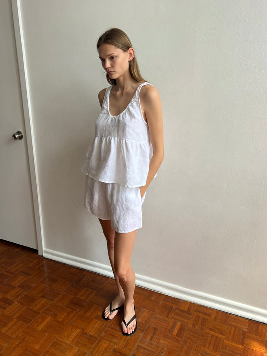 A woman stands indoors on a wooden floor, wearing the Gathered Linen Set - White with black flip-flops, beside a light-colored wall and a closed door.