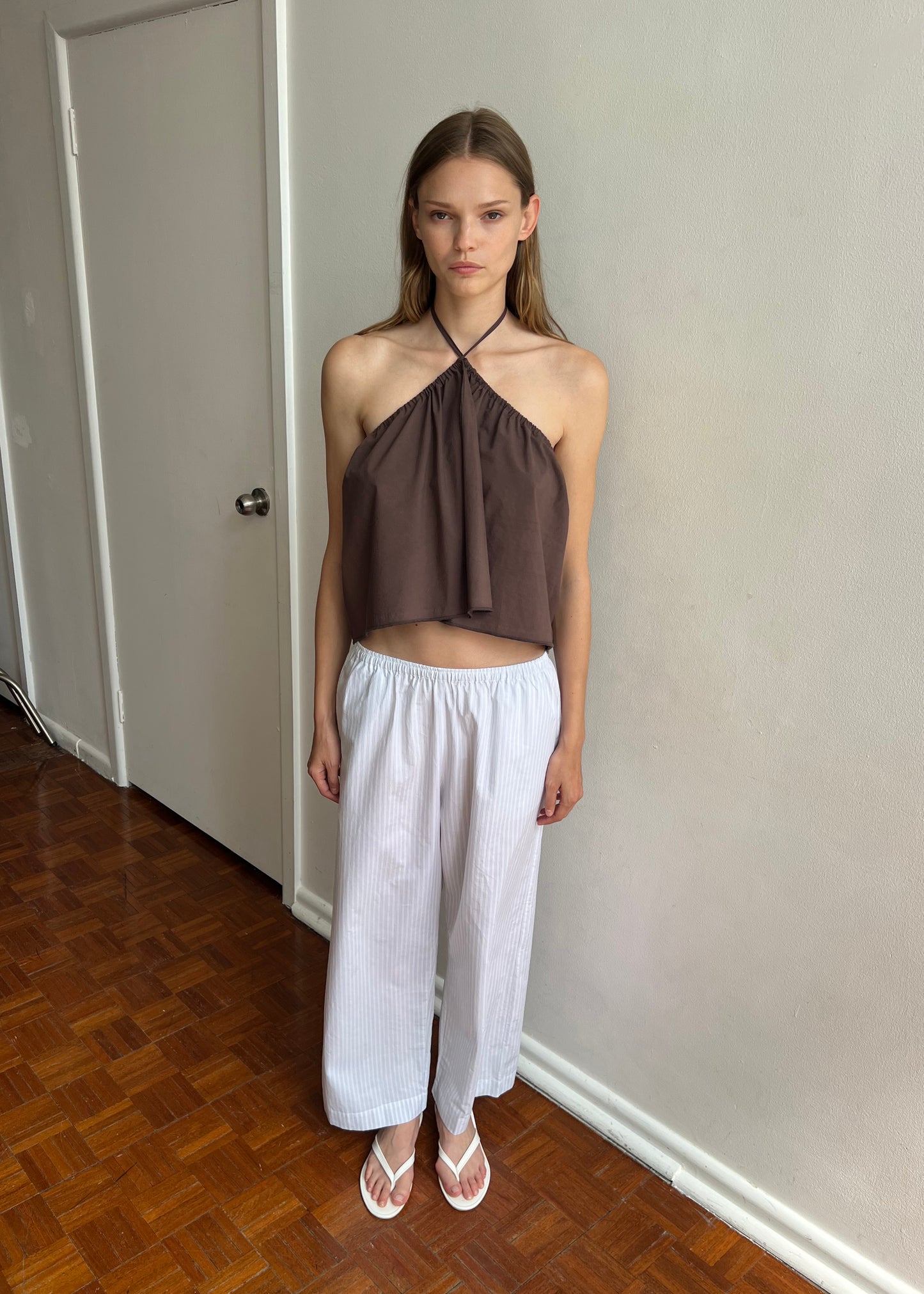 A woman with straight, light brown hair wears a brown halter top and Deiji Studios' Ease Trouser in Sage Stripe with white sandals. She stands indoors on wooden flooring near an off-white wall and a closed door, looking neutral.