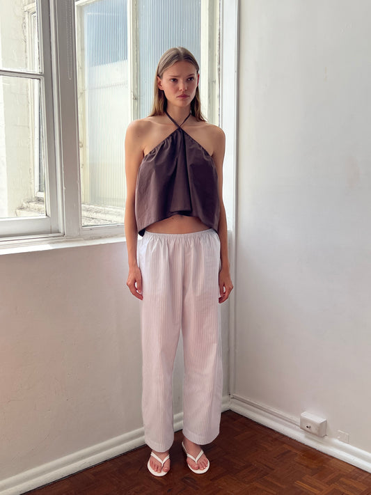 A woman stands by a sunlit window, wearing a brown halter-style top and Deiji Studios Ease Trouser in Sage Stripe. She has straight, shoulder-length hair and a neutral expression. The room features white walls and a wooden floor.