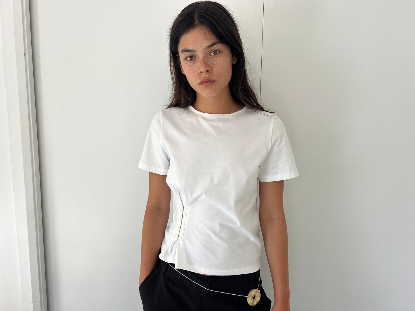 A young woman with long dark hair and freckles stands against a white wall, wearing the Pinch Tee - White by Deiji Studios and black pants, one hand in her pocket and a neutral expression.
