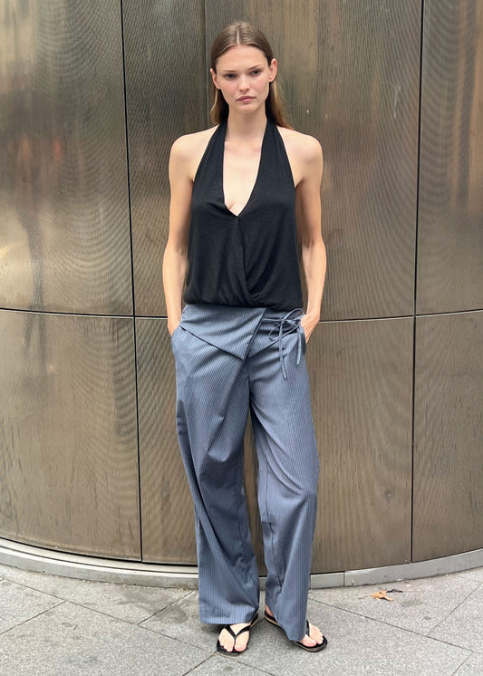 A woman stands outdoors by a metallic wall, wearing a black halter top and the Folded Wool Pant - Navy Stripe with her hands in her pockets and a neutral expression.