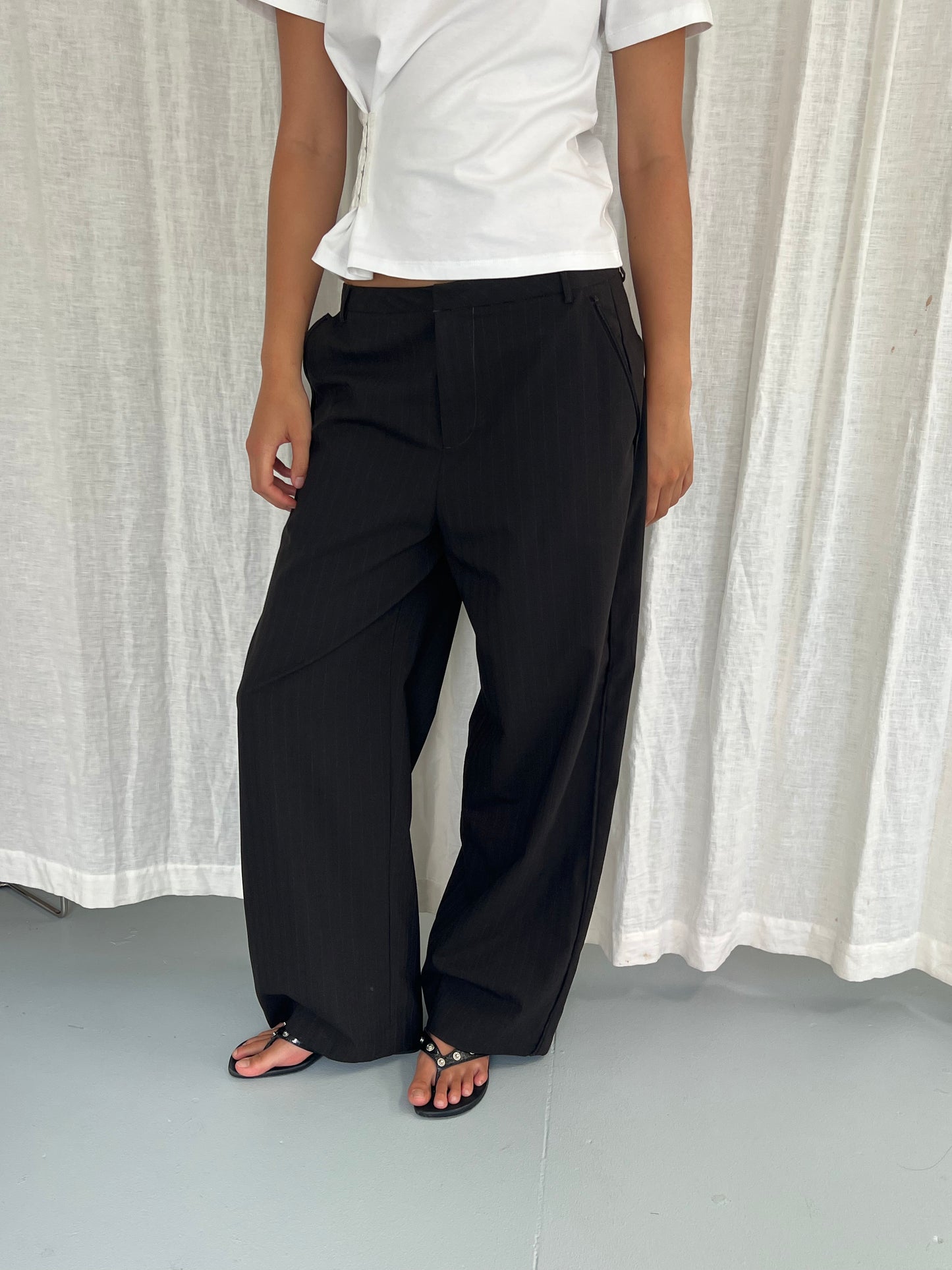 A person stands before a white curtain, wearing a white short-sleeve top, Deiji Studios Bind Pant in Charred Black Stripe, and black sandals. Their face is out of frame.