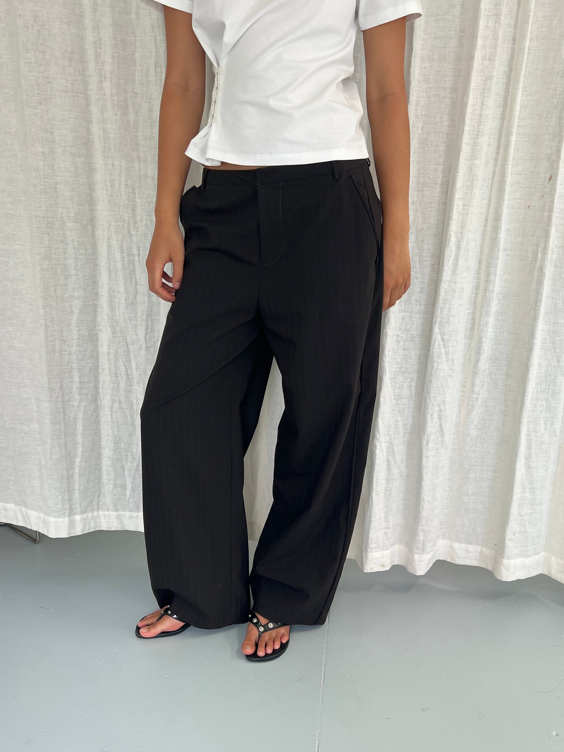 A person stands before a white curtain, wearing a white short-sleeve top, Deiji Studios Bind Pant in Charred Black Stripe, and black sandals. Their face is out of frame.