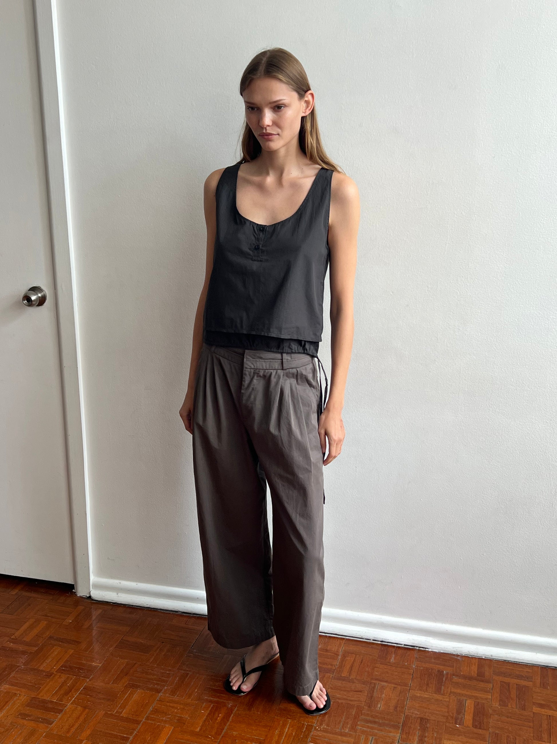 A woman with straight light brown hair and a neutral expression stands indoors against a white wall, wearing Deiji Studios' Layered Top - Black Stripe, loose gray pants, and black sandals on a brown wooden floor.