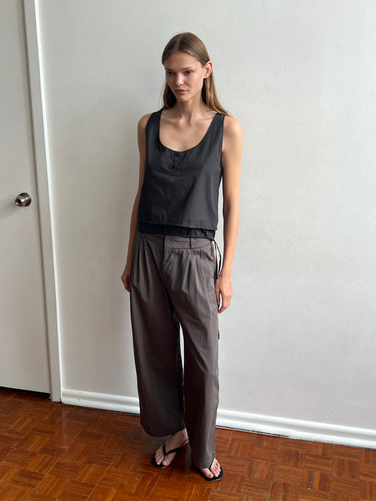 A woman with straight light brown hair and a neutral expression stands indoors against a white wall, wearing Deiji Studios' Layered Top - Black Stripe, loose gray pants, and black sandals on a brown wooden floor.