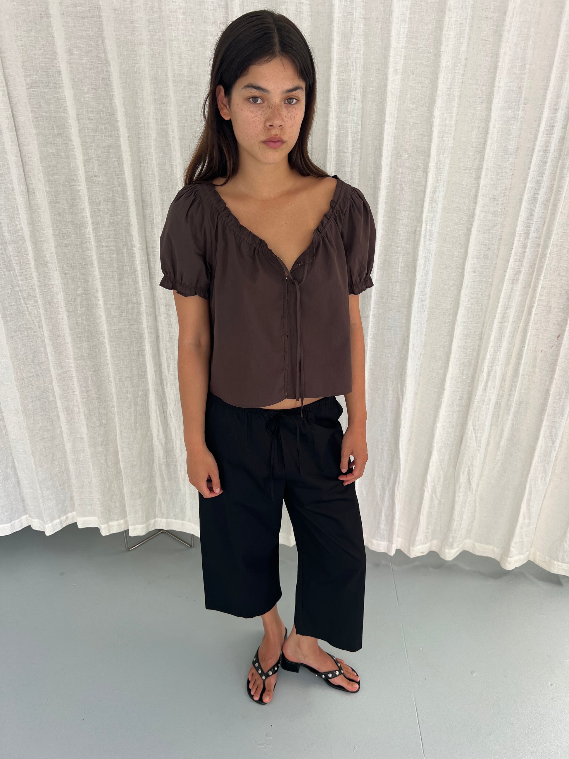 A young woman with long dark hair stands indoors on a light grey floor before white curtains, wearing a Deiji Studios Close Shirt in Sepia, black wide-leg pants, and black sandals. She gazes slightly to the side with a neutral expression.
