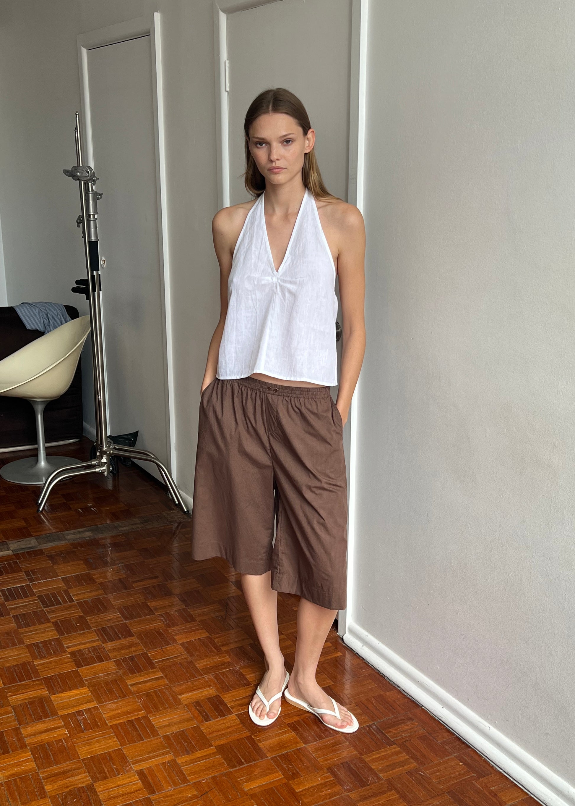 A person stands indoors on a wooden floor wearing Deiji Studios' Long Lounger - Carob Check, styled as knee-length shorts in organic cotton, paired with a sleeveless white halter top and white sandals. Their hands are in their pockets and they have straight hair.