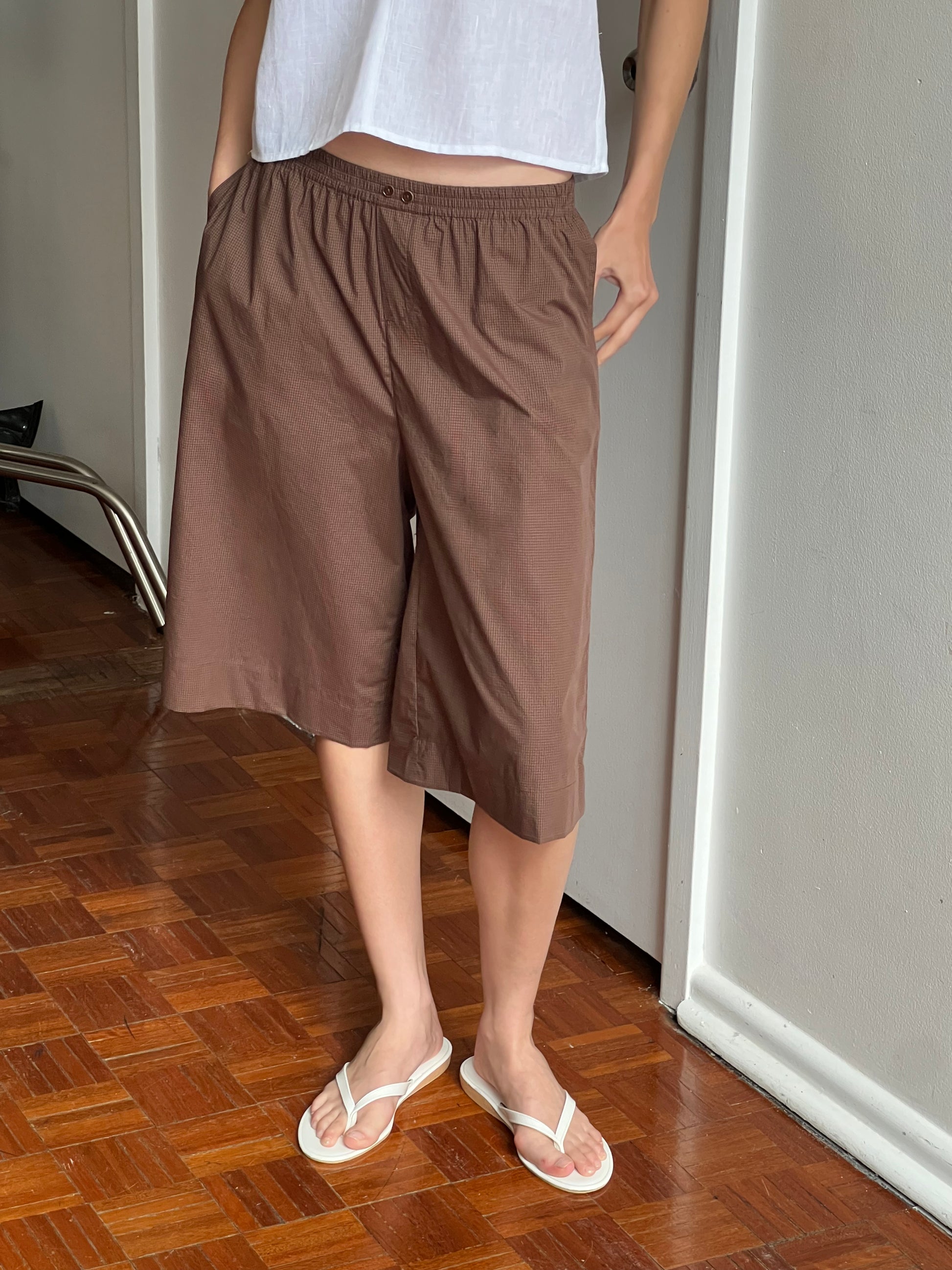 Person wearing the Long Lounger - Carob Check, brown knee-length shorts, and white flip-flops stands indoors on a wooden floor by a white wall, hands in pockets—classic comfort in organic cotton.
