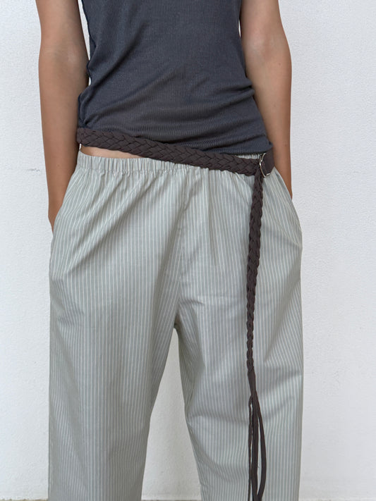 A person stands against a white background with hands in their pockets, wearing a dark sleeveless top, light striped pants, and the 0 Plaited Belt - Chocolate—crafted from soft organic cotton with tassels.