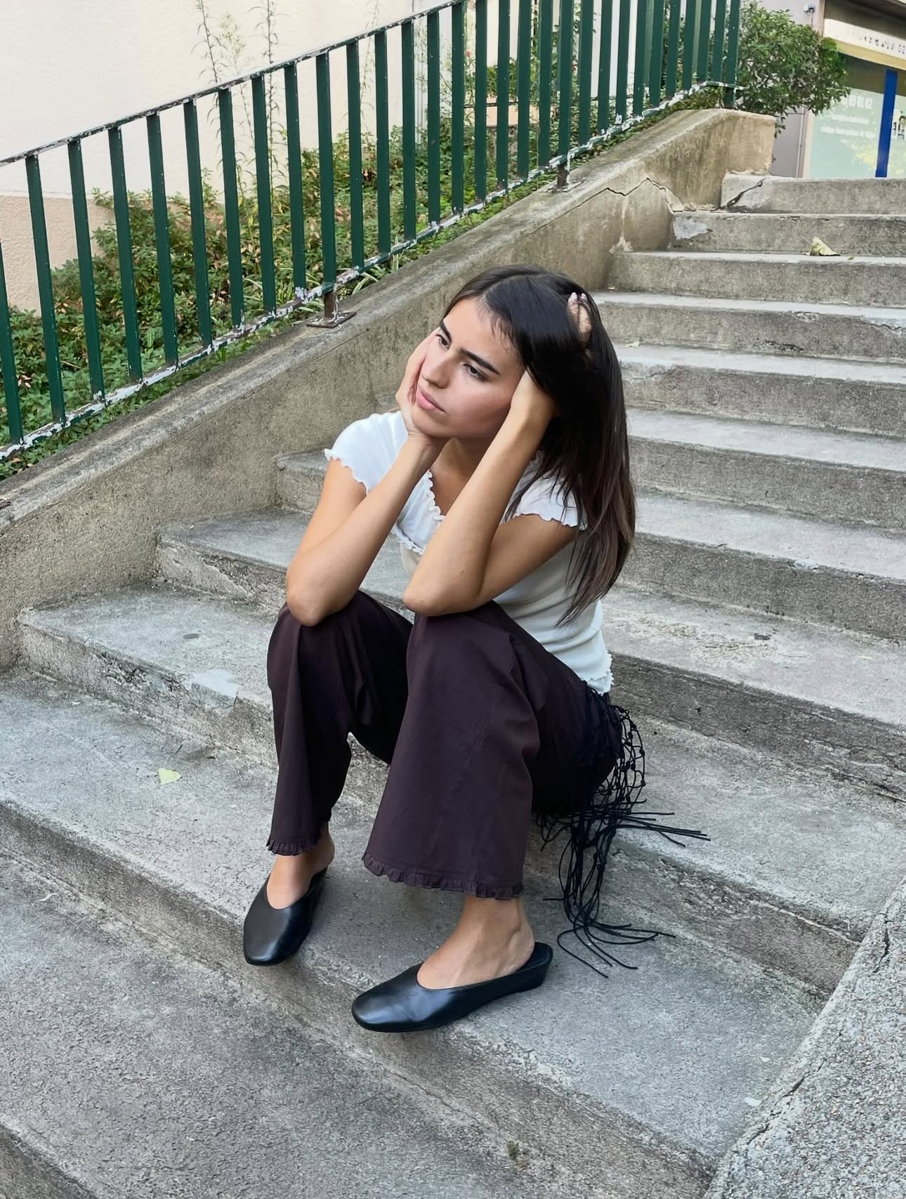 A young woman with long dark hair sits on outdoor steps, resting her head in her hands and gazing thoughtfully to the side. She wears a white top, black flats, and the Flounce Pant in a dark shade with a wide-leg silhouette.