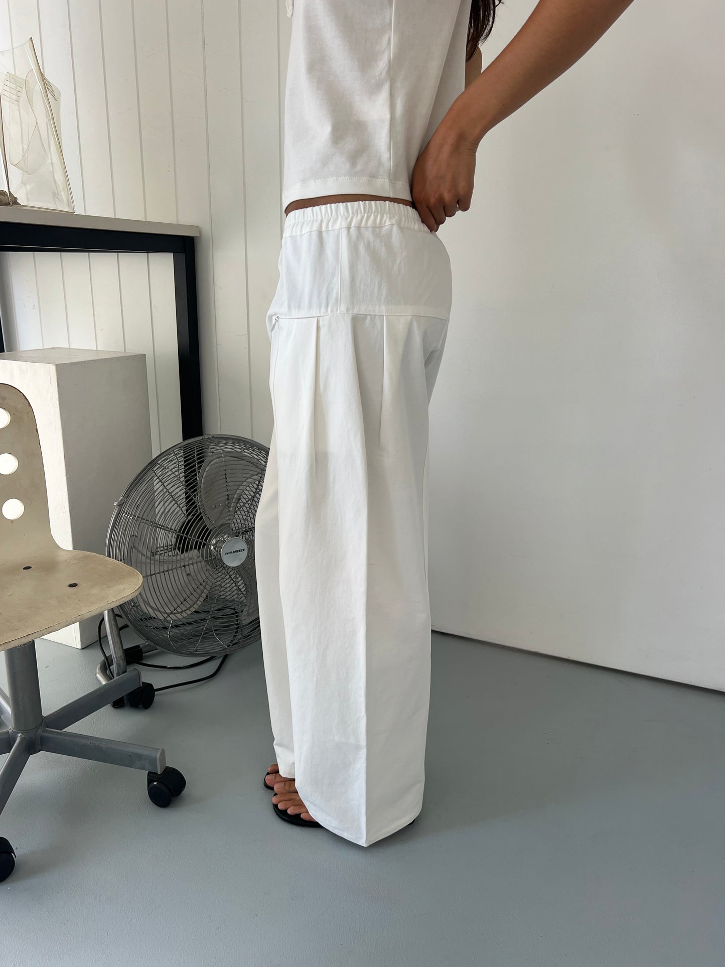 Someone stands indoors in a minimalist room, adjusting the fit of the Pleat Pant - White with an elastic waistband and loose pleats. Only part of their upper body is visible, along with a fan, chair, and desk in the background.