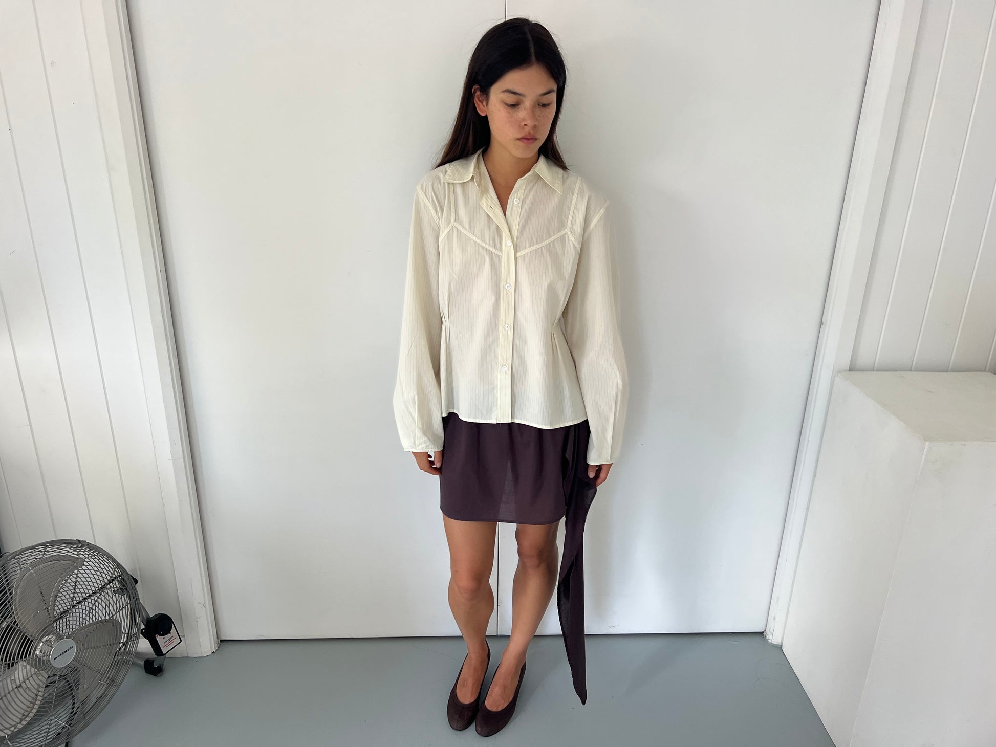A woman with long dark hair stands indoors against a white wall, wearing the Cami Overshirt - Citrus Stripe and a dark brown asymmetrical hem skirt with brown shoes. A silver floor fan is visible on the left as she looks down.