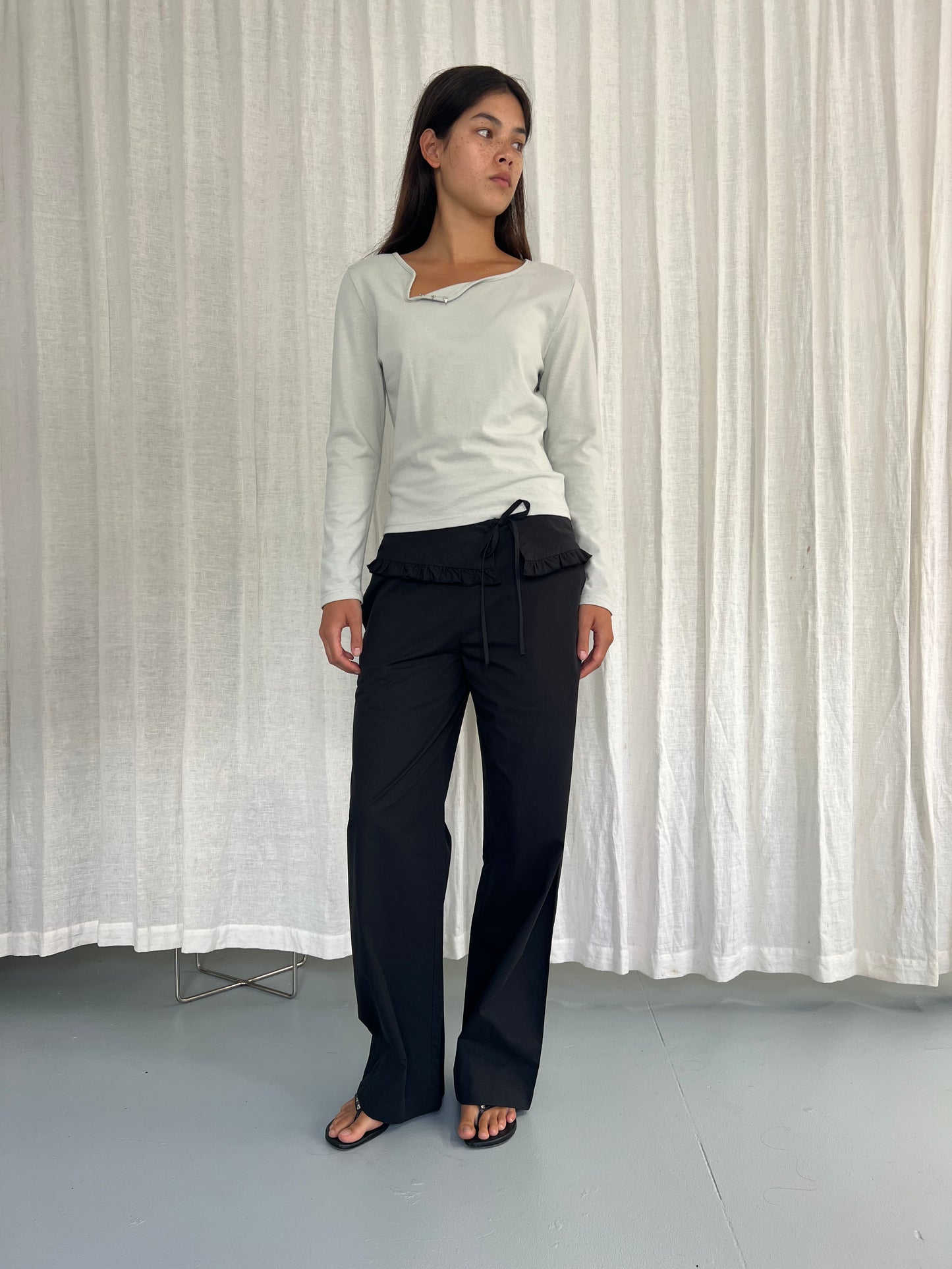 A woman stands indoors by white curtains, wearing a white long-sleeve top, the Flounce Belt Pant - Black with ruffle detail, and black flip-flops, looking slightly left.
