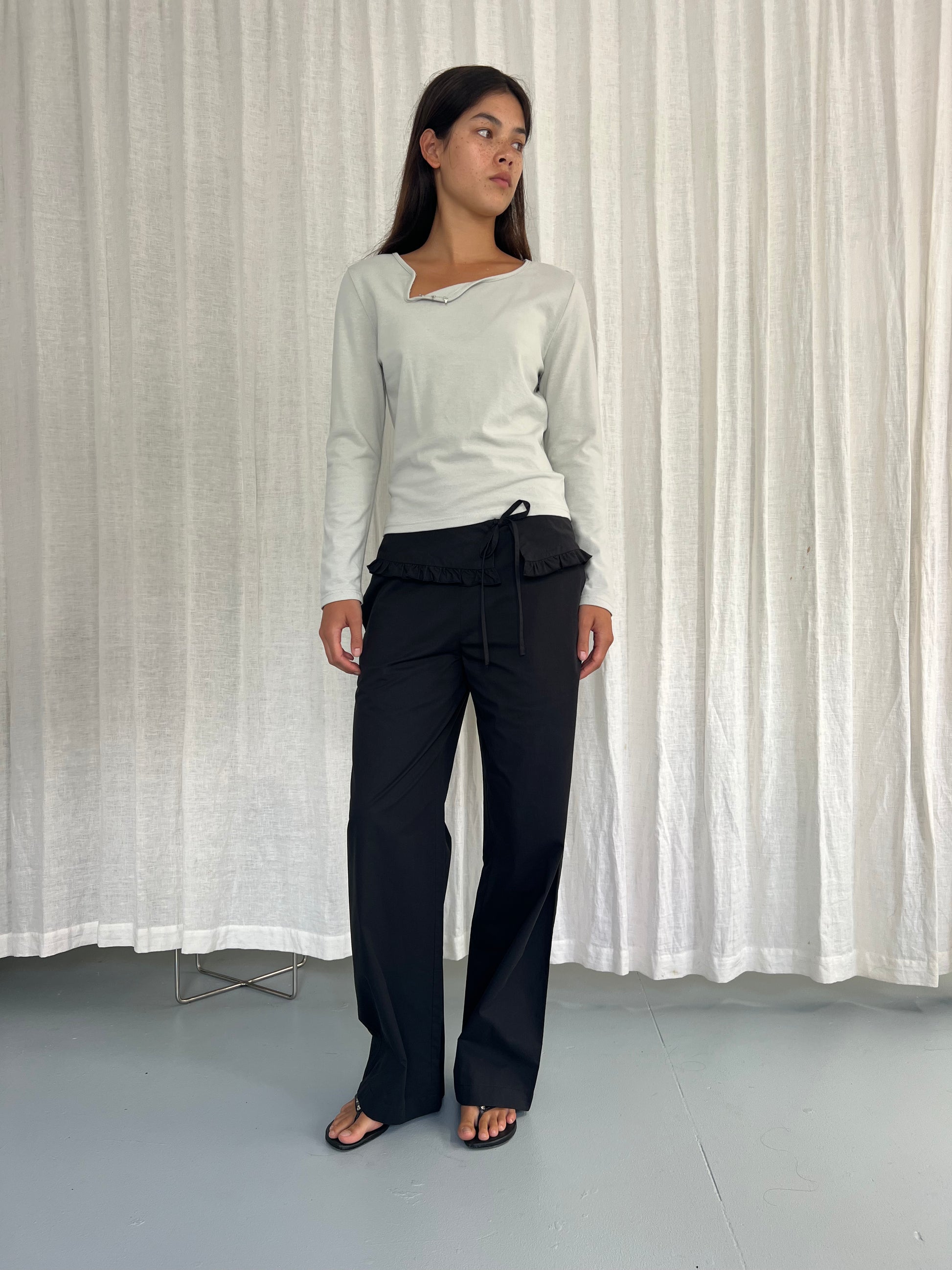 A woman stands indoors by white curtains, wearing a white long-sleeve top, the Flounce Belt Pant - Black with ruffle detail, and black flip-flops, looking slightly left.