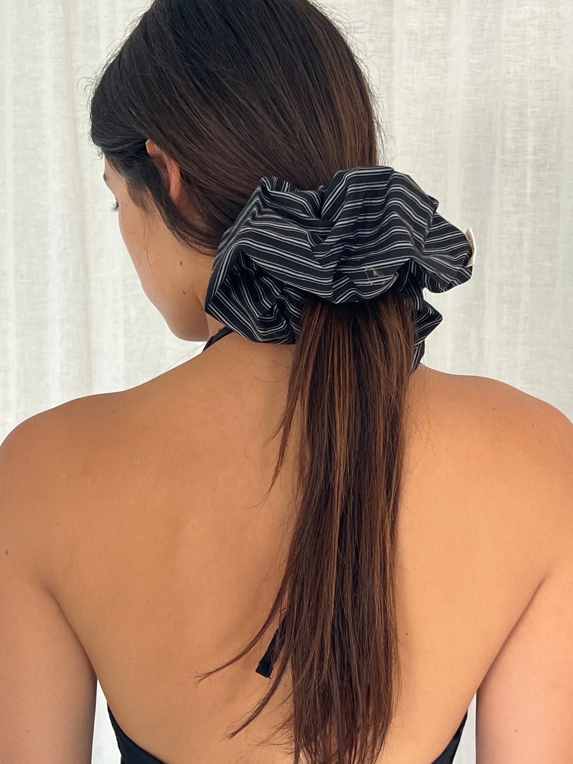 A woman with long brown hair, seen from behind, wears the Mono Stripe scrunchie and a black strapless top against a light textured background.