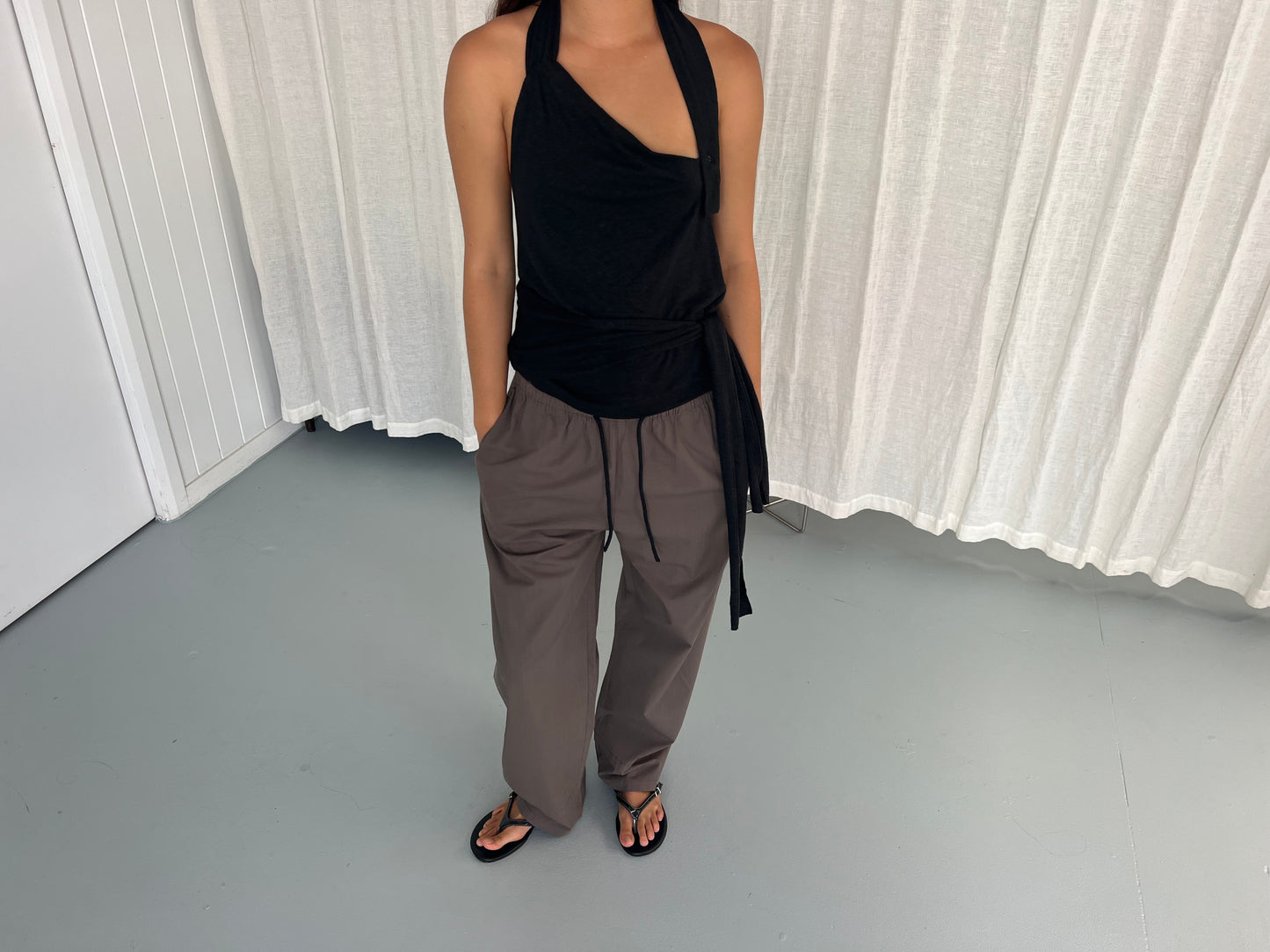 A person wearing the Drape Halter - Black from Deiji Studios, loose gray drawstring pants, and black sandals stands indoors on a light gray floor in front of white curtains. Their face is not visible.