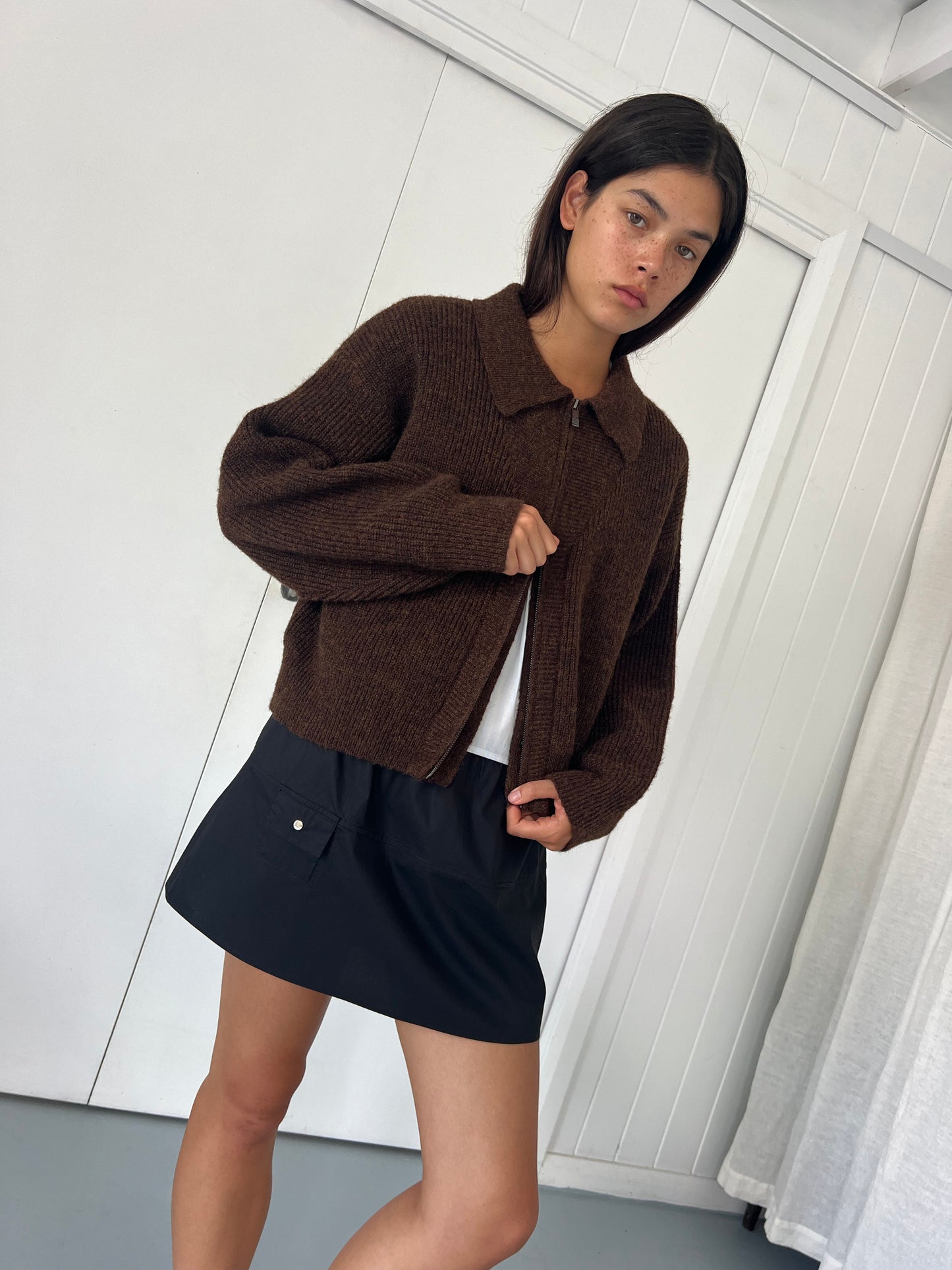 A person with straight dark hair wears the Double Layer Cardigan - Brown by Deiji Studios over a white top and black skirt, standing indoors against a white paneled wall and looking at the camera.