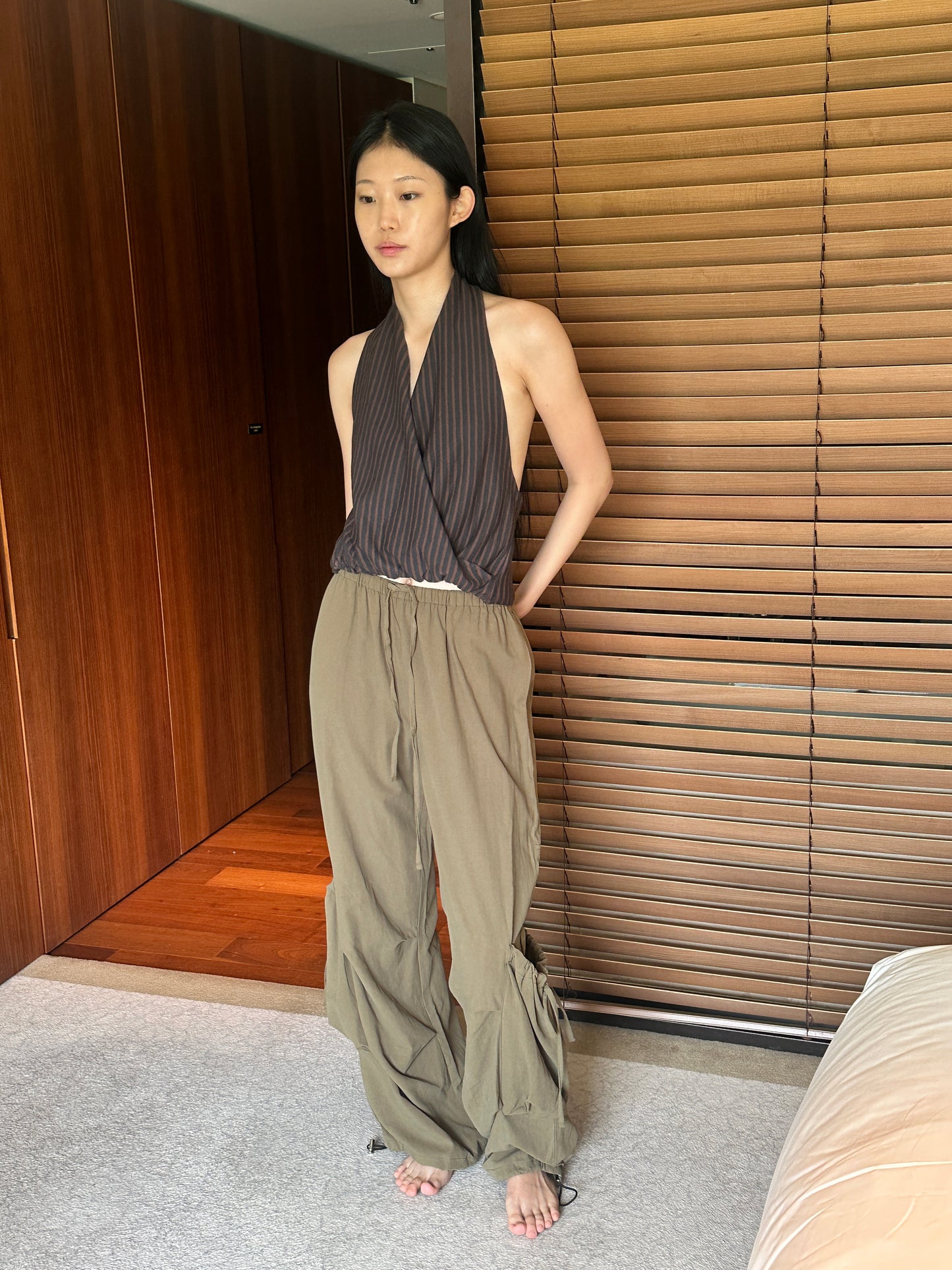 A woman with long dark hair stands in a bedroom wearing the Wrap Halter Top - Brown Grey Stripe by Deiji Studios, a sheer brown and grey striped halter top with an open back, and khaki drawcord trousers.