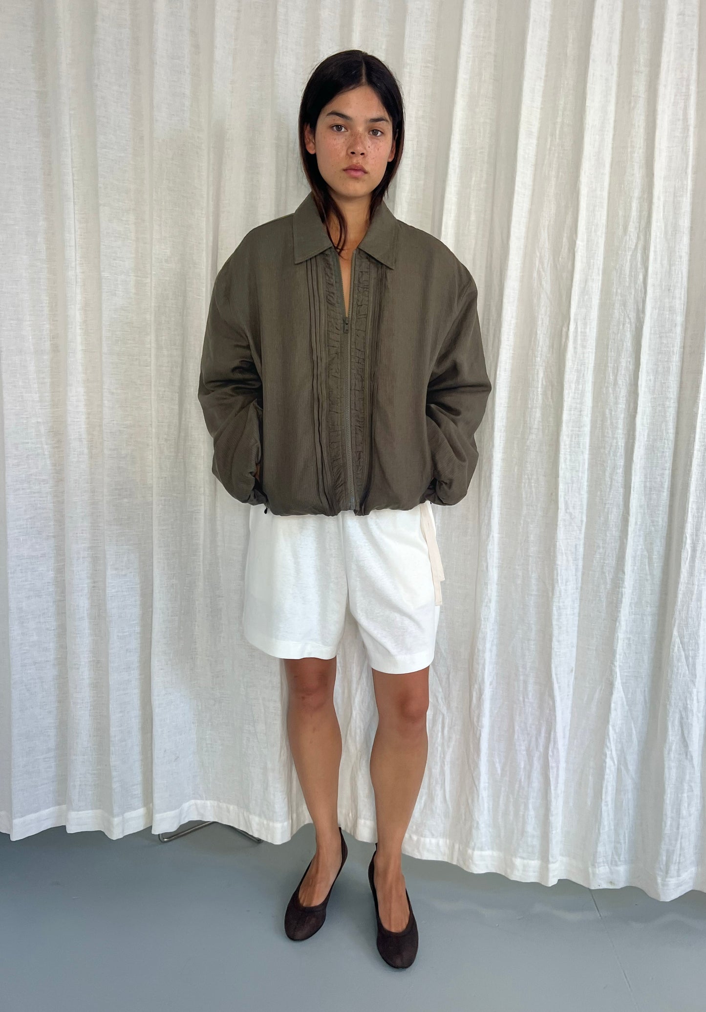 A person stands before a white curtain, wearing the Pintuck Jacket - Khaki Stripe from Deiji Studios, paired with white shorts and black flats. Their hands are in their pockets and their expression is neutral.