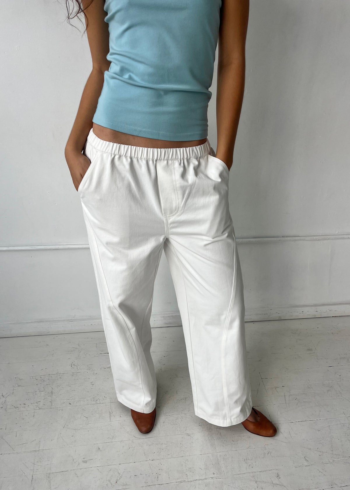 Cropped to the lower body, a woman stands against a white wall with hands in the pockets of the Curve Pant - White by Deiji Studios, a softly structured pant with a relaxed fit and side pockets, styled with the Scallop Tank in Sky.