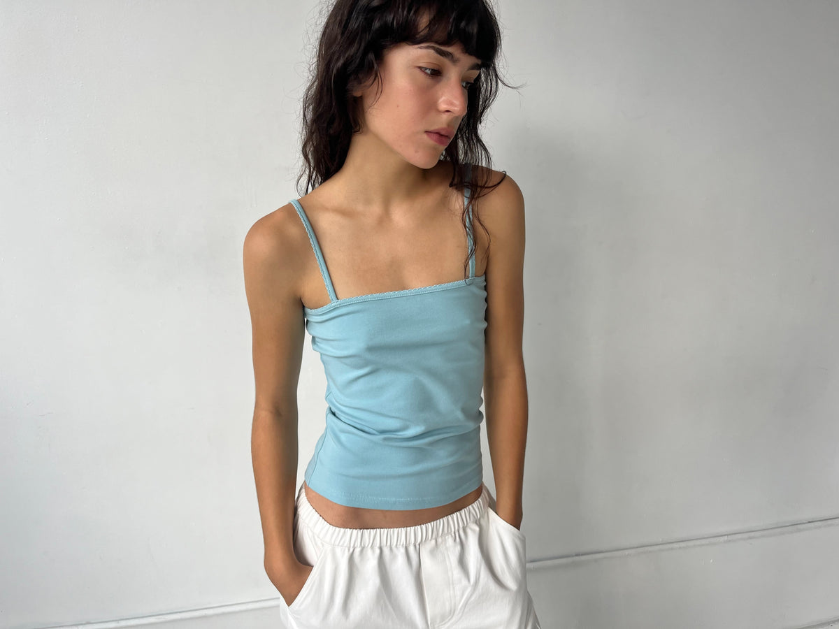 A woman stands against a white wall with hands in her pockets, wearing the Scallop Tank - Sky by Deiji Studios, a fitted ribbed tank with fine straps and scallop trim, styled with the Curve Pant in White.