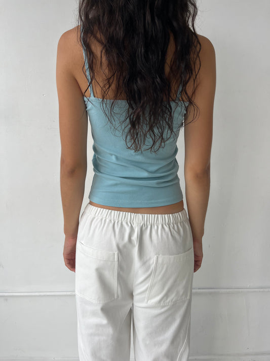 A woman stands facing a white wall, seen from behind, wearing the Scallop Tank - Sky by Deiji Studios, a fitted ribbed tank with fine straps and scallop trim along the straps and back neckline, styled with the Curve Pant in White.