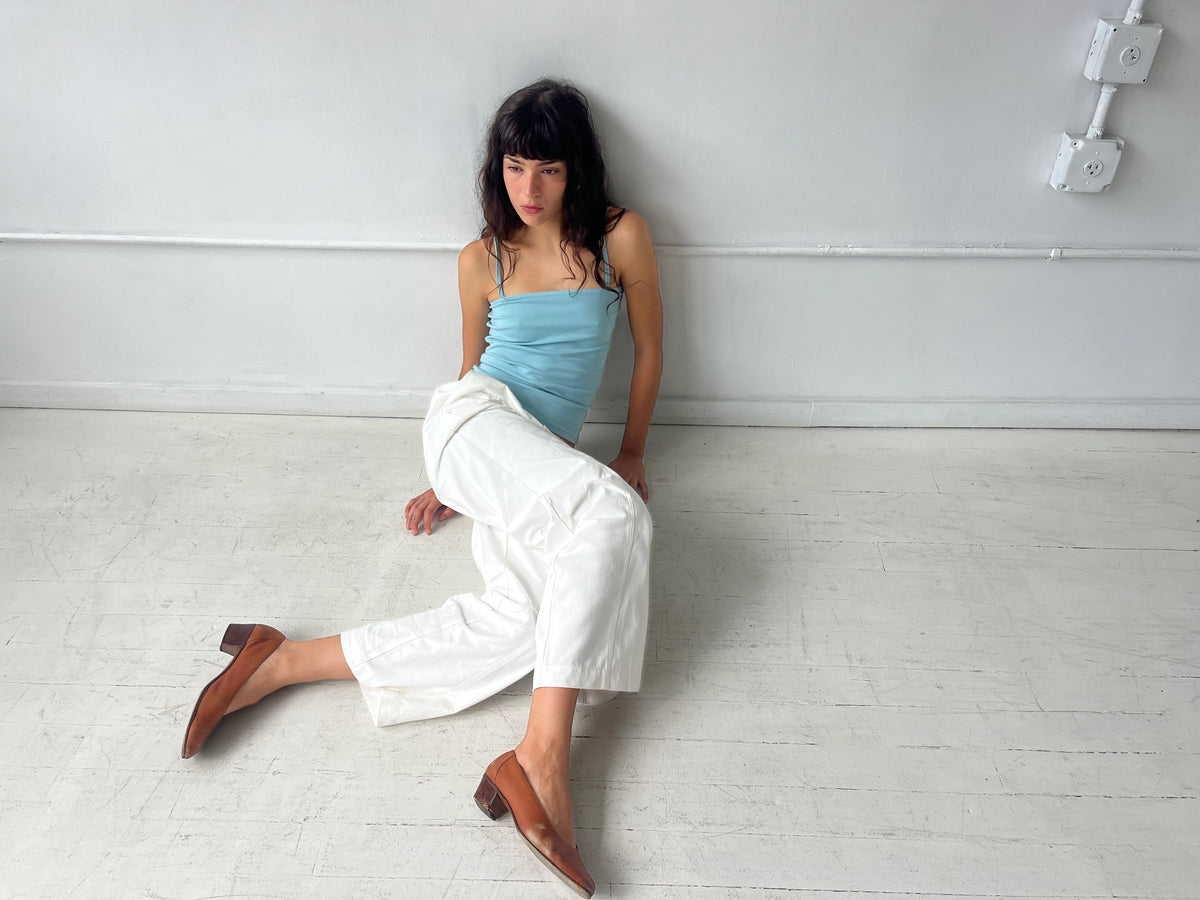 A woman sits on a white studio floor, wearing the Curve Pant - White by Deiji Studios, a softly structured pant with a relaxed fit and curved leg, styled with the Scallop Tank in Sky and brown heels.