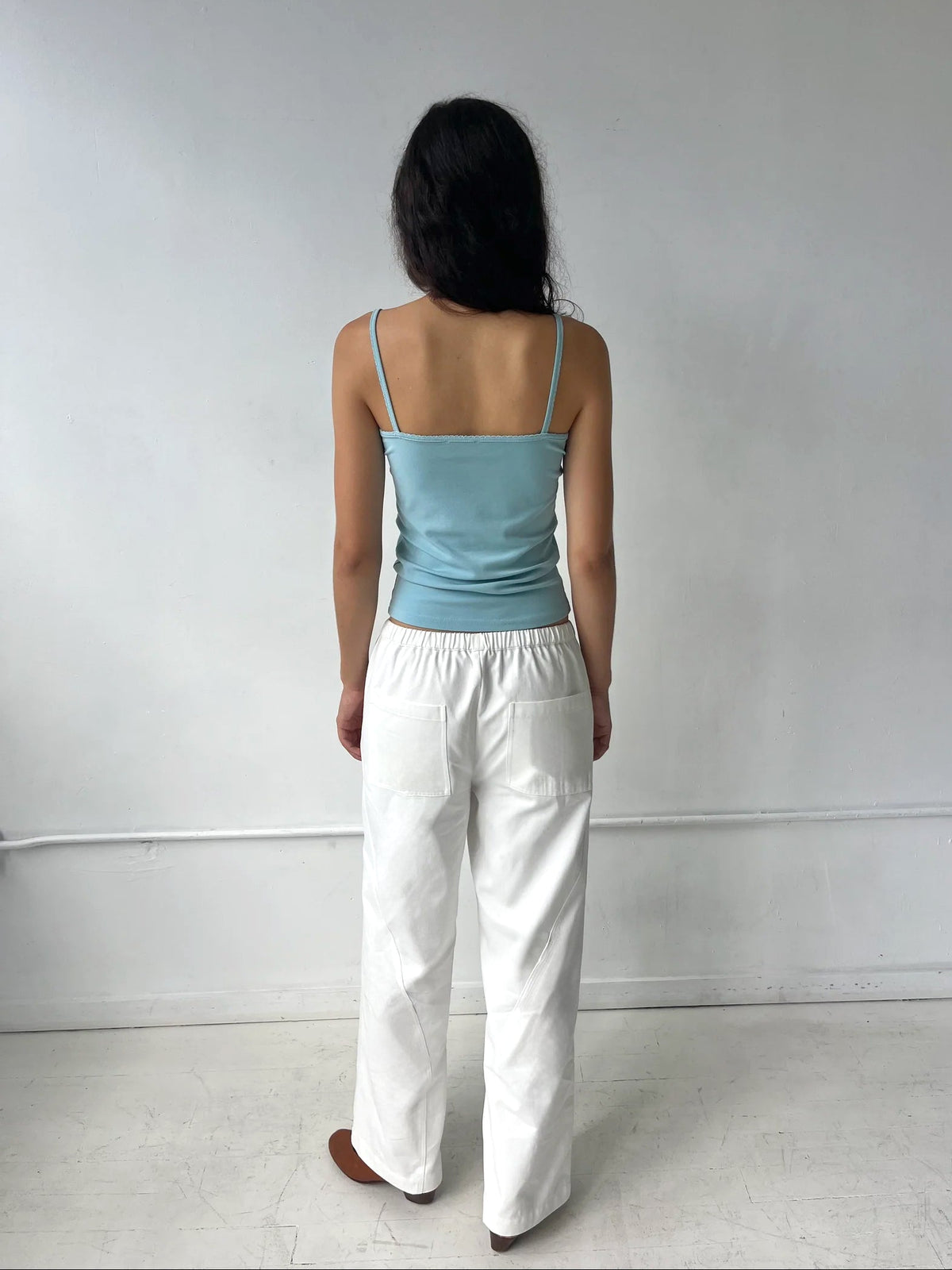 A woman stands facing a white wall, seen from behind, wearing the Curve Pant - White by Deiji Studios, a softly structured pant with a relaxed fit and back patch pockets, styled with the Scallop Tank in Sky.