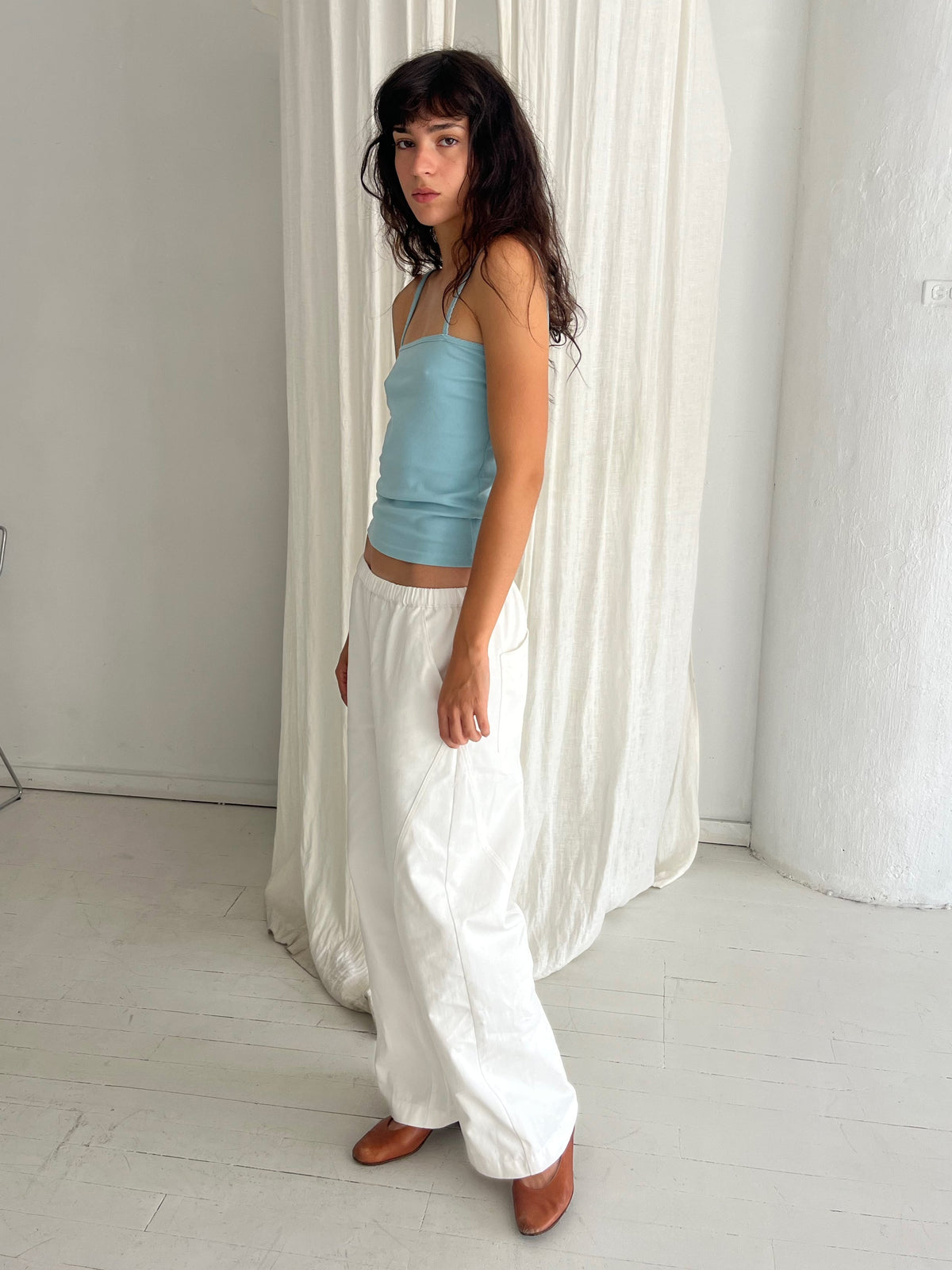 A woman with dark wavy hair stands in profile against a white curtain, wearing the Curve Pant - White by Deiji Studios, a softly structured pant with a relaxed fit and subtle curved leg, styled with the Scallop Tank in Sky.