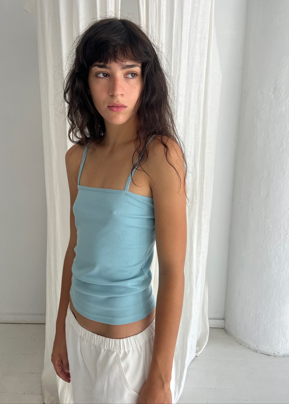 A woman with dark wavy hair stands against a white curtain, wearing the Scallop Tank - Sky by Deiji Studios, a fitted ribbed tank with fine straps and scallop trim, styled with the Curve Pant in White.