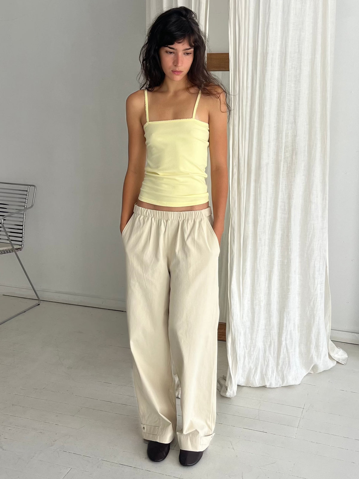 A woman stands against a white curtain with hands in her pockets, wearing the Snap Tab Pant - Stone by Deiji Studios, a relaxed wide-leg pant with an elasticated waistband and snap tabs at the hem, styled with the Scallop Tank in Lemon.