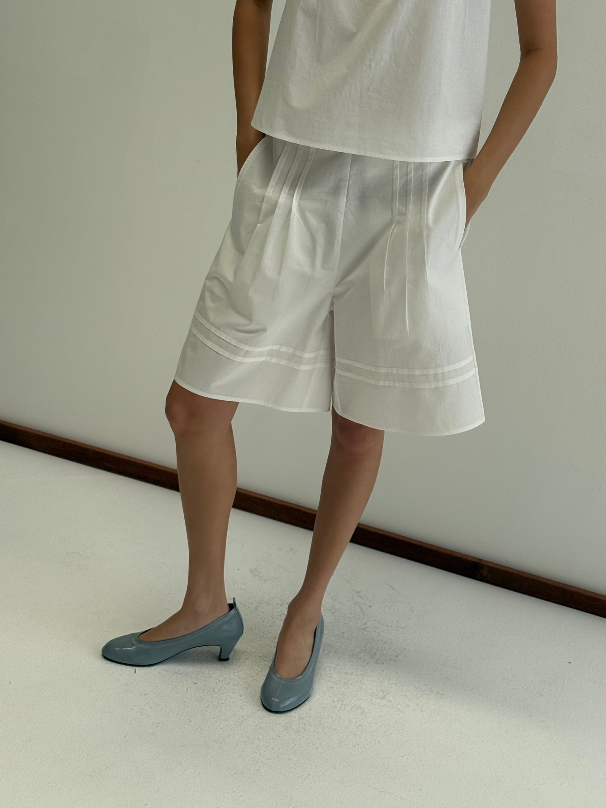 A person stands against a plain wall wearing Deiji Studios' Pintuck Short in white paired with a loose white top. Light blue low-heeled shoes complete the look, hands in pockets and head out of frame.