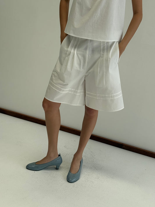 A person stands against a plain wall wearing Deiji Studios' Pintuck Short in white paired with a loose white top. Light blue low-heeled shoes complete the look, hands in pockets and head out of frame.