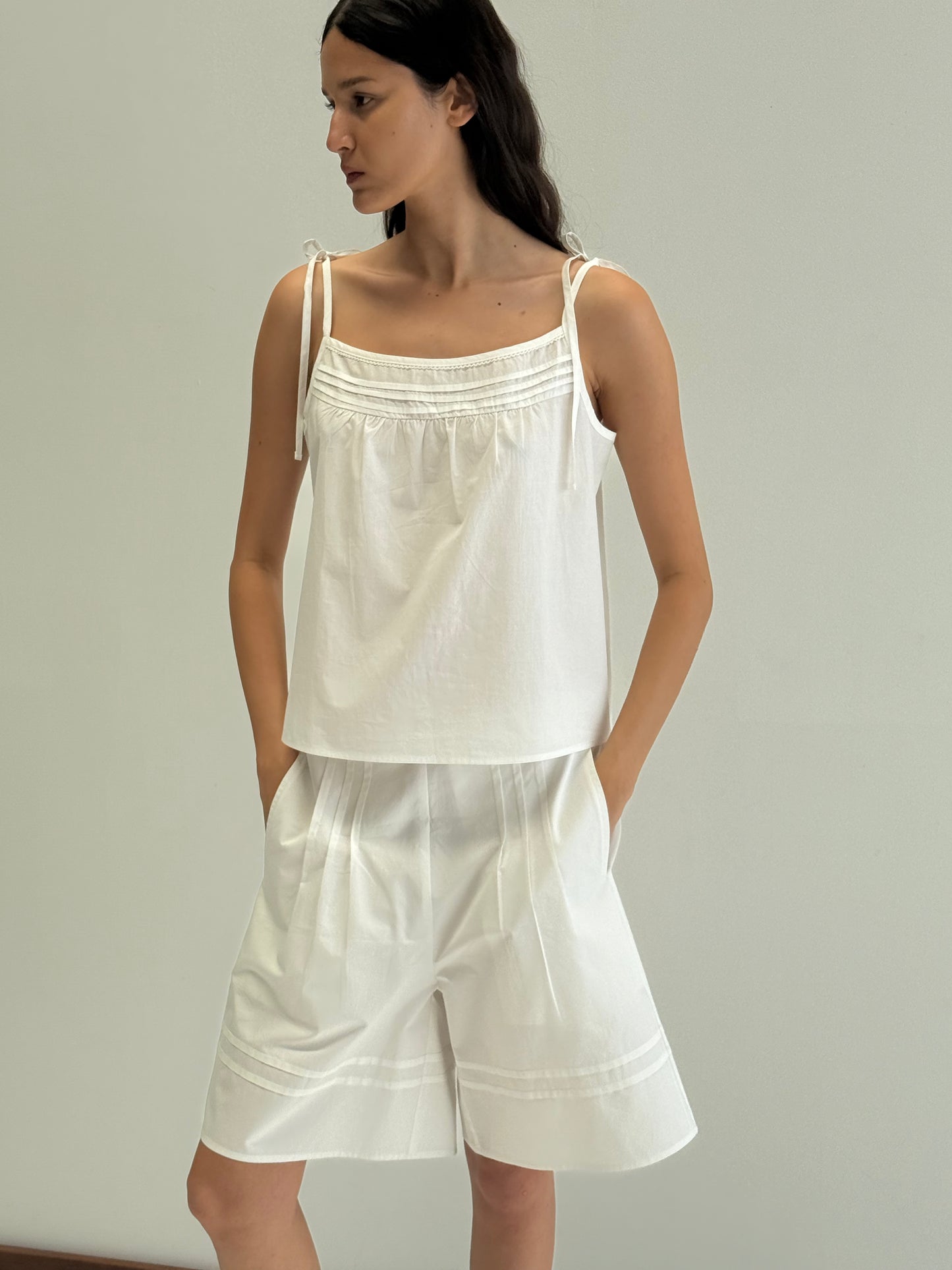 A woman with long dark hair stands against a plain light background, wearing Deiji Studios' Tied Cami - White with matching loose white shorts. She looks to the side, her hands in her pockets.