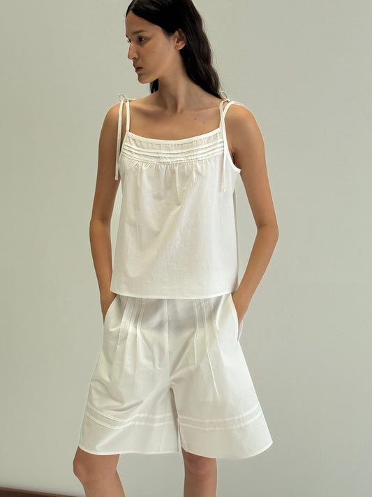 A woman with long dark hair stands against a plain light background, wearing Deiji Studios' Tied Cami - White with matching loose white shorts. She looks to the side, her hands in her pockets.