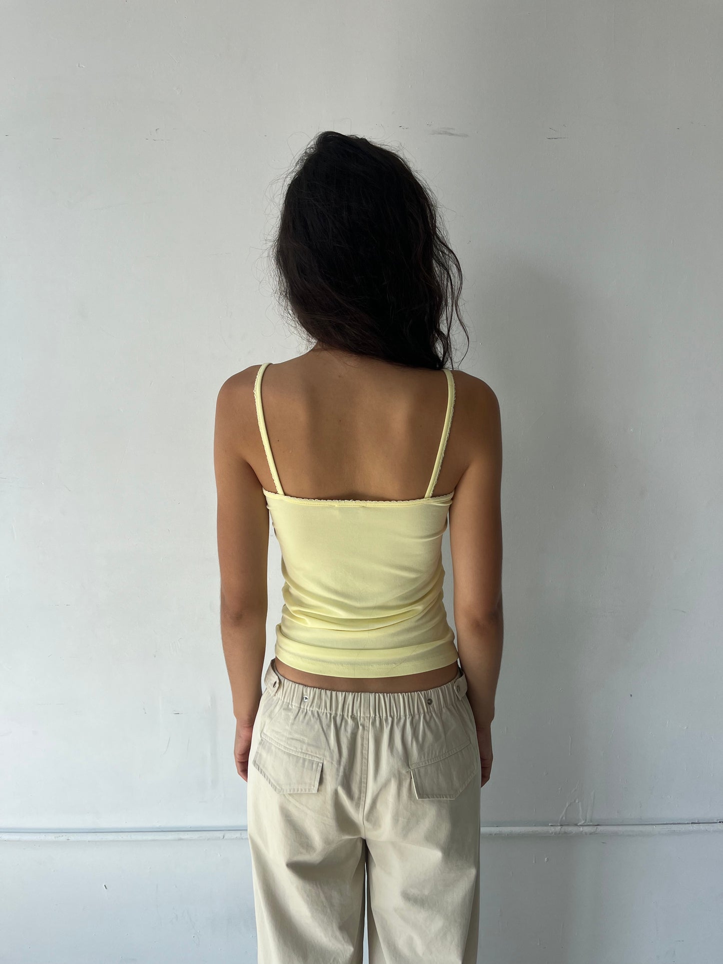 A woman stands facing a white wall, seen from behind, wearing the Scallop Tank - Lemon by Deiji Studios, a fitted ribbed tank with fine straps and scallop trim along the straps and back neckline, with cream wide-leg pants.