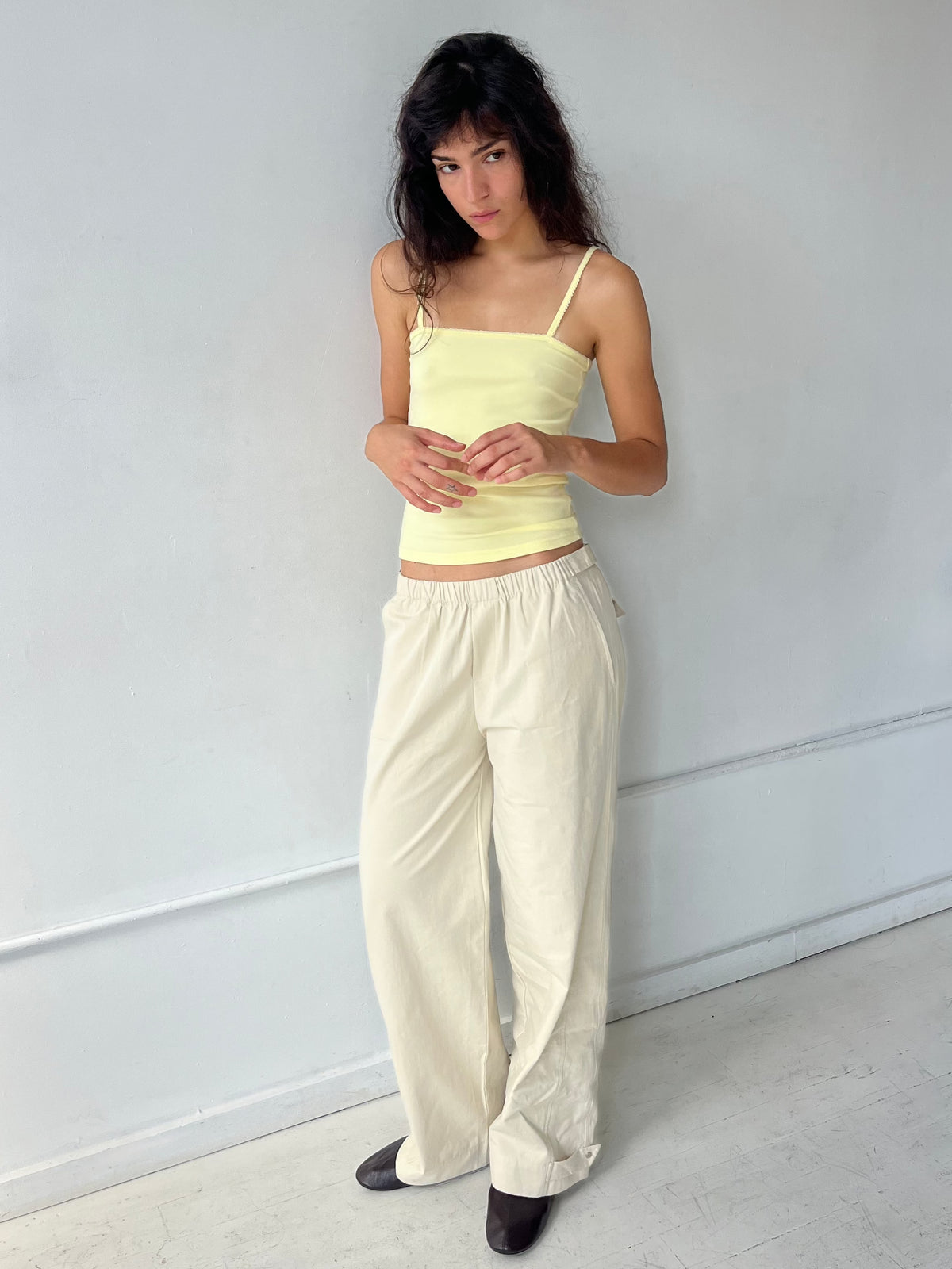 A woman with dark wavy hair stands against a white wall, wearing the Snap Tab Pant - Stone by Deiji Studios, a relaxed wide-leg pant with an elasticated waistband and snap tabs at the hem, styled with the Scallop Tank in Lemon.