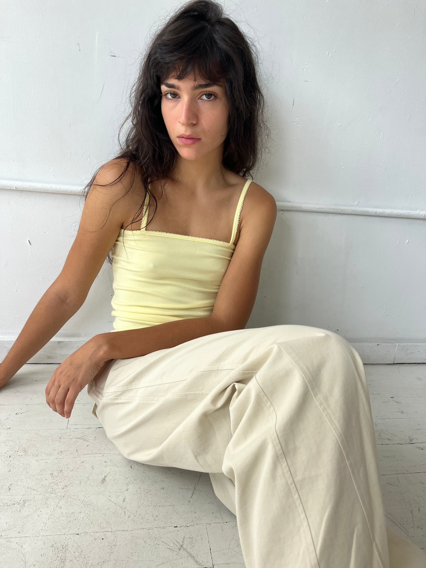 A woman sits on a white studio floor with legs extended, wearing the Scallop Tank - Lemon by Deiji Studios, a fitted ribbed tank with fine straps and scallop trim, with cream wide-leg pants.