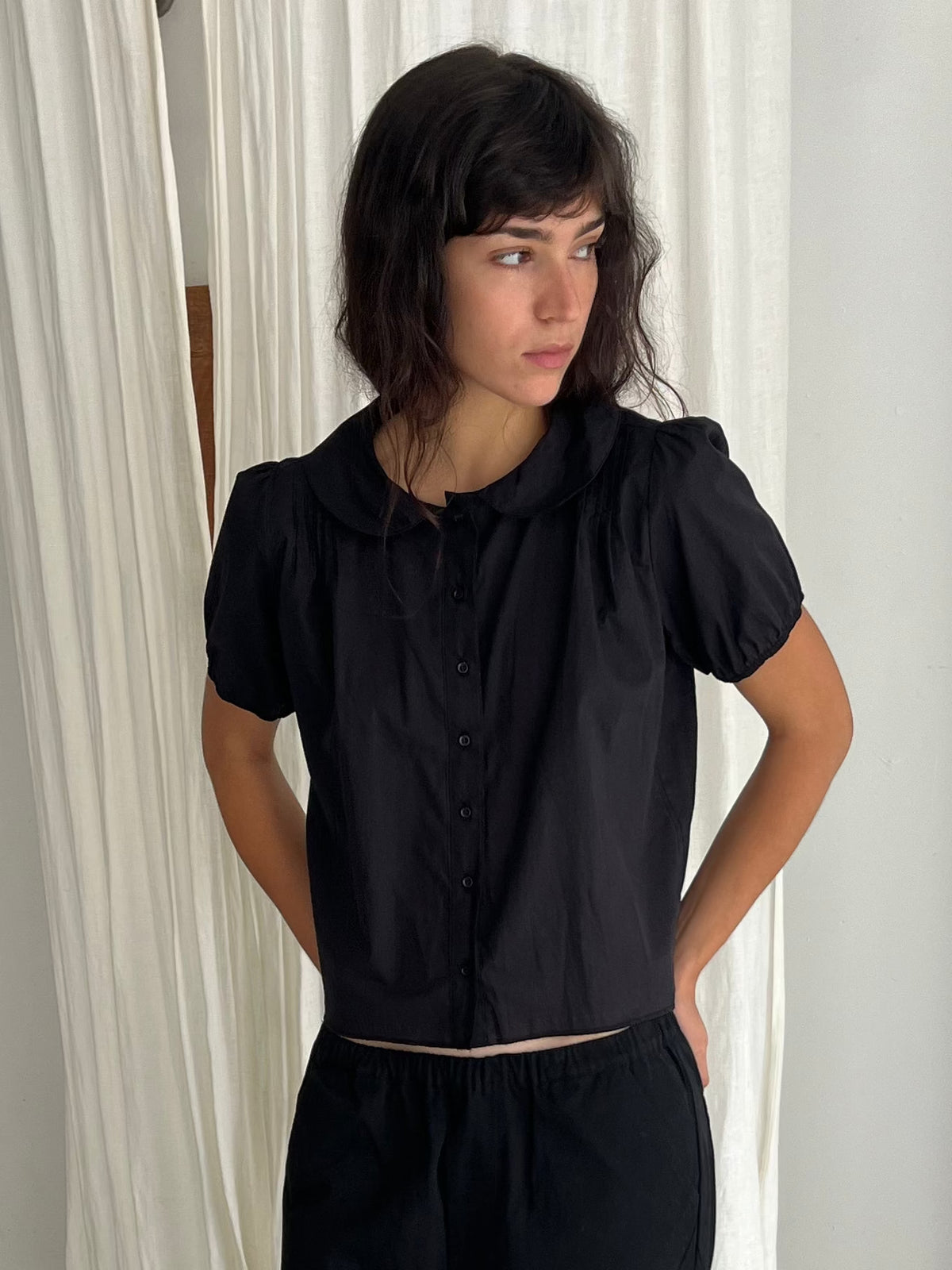 A woman with dark wavy hair stands against a white curtain, looking to the side, wearing the Petal Top - Black by Deiji Studios, a lightweight cotton voile top with a rounded peter pan collar, button-through front and puffed short sleeves.