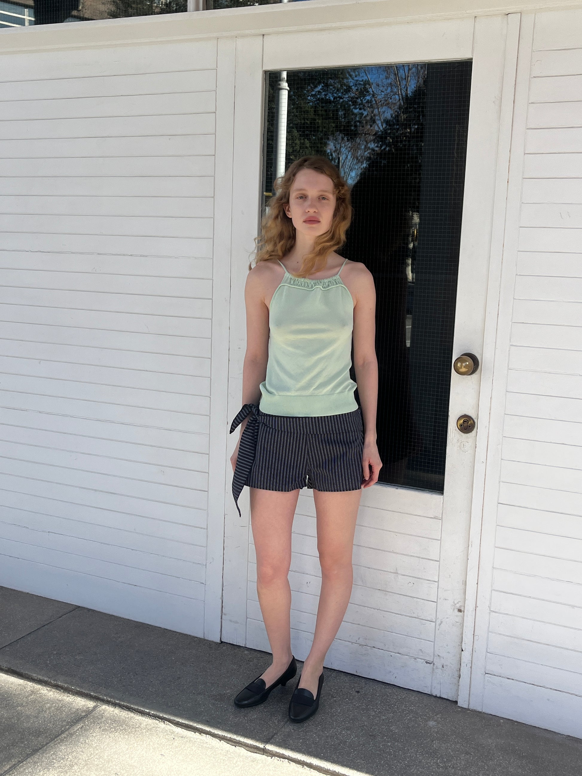 A woman with wavy blonde hair stands before a white door on a sunny day, wearing the mint Drawcord Knit Top by Deiji Studios, navy striped side-tie shorts, and black loafers.