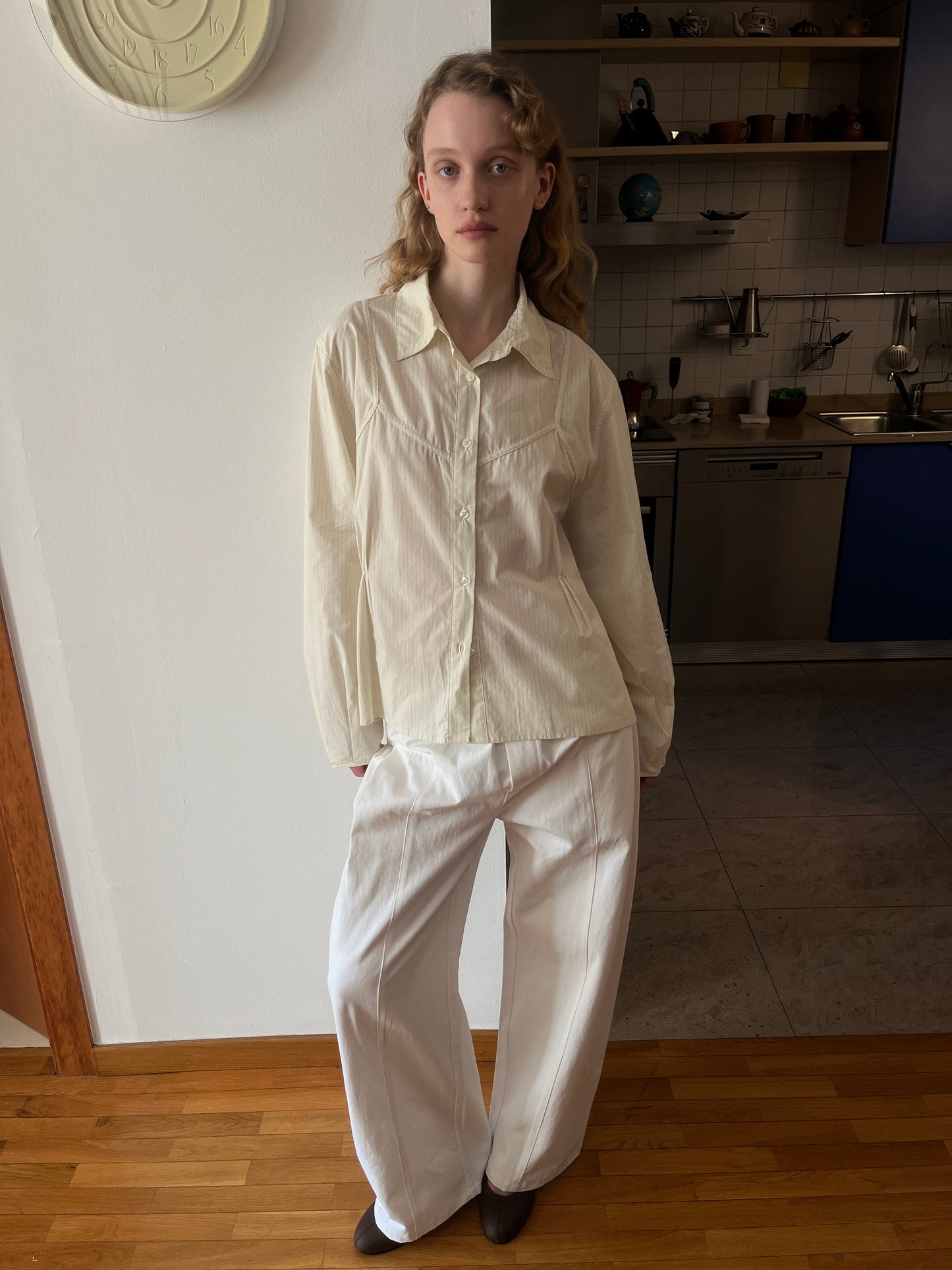 A person with wavy blonde hair stands indoors, wearing a loose-fitting cream button-up shirt and the Pleat Pant - White by Deiji Studios. The background features a modern kitchen with blue cabinets and shelves.