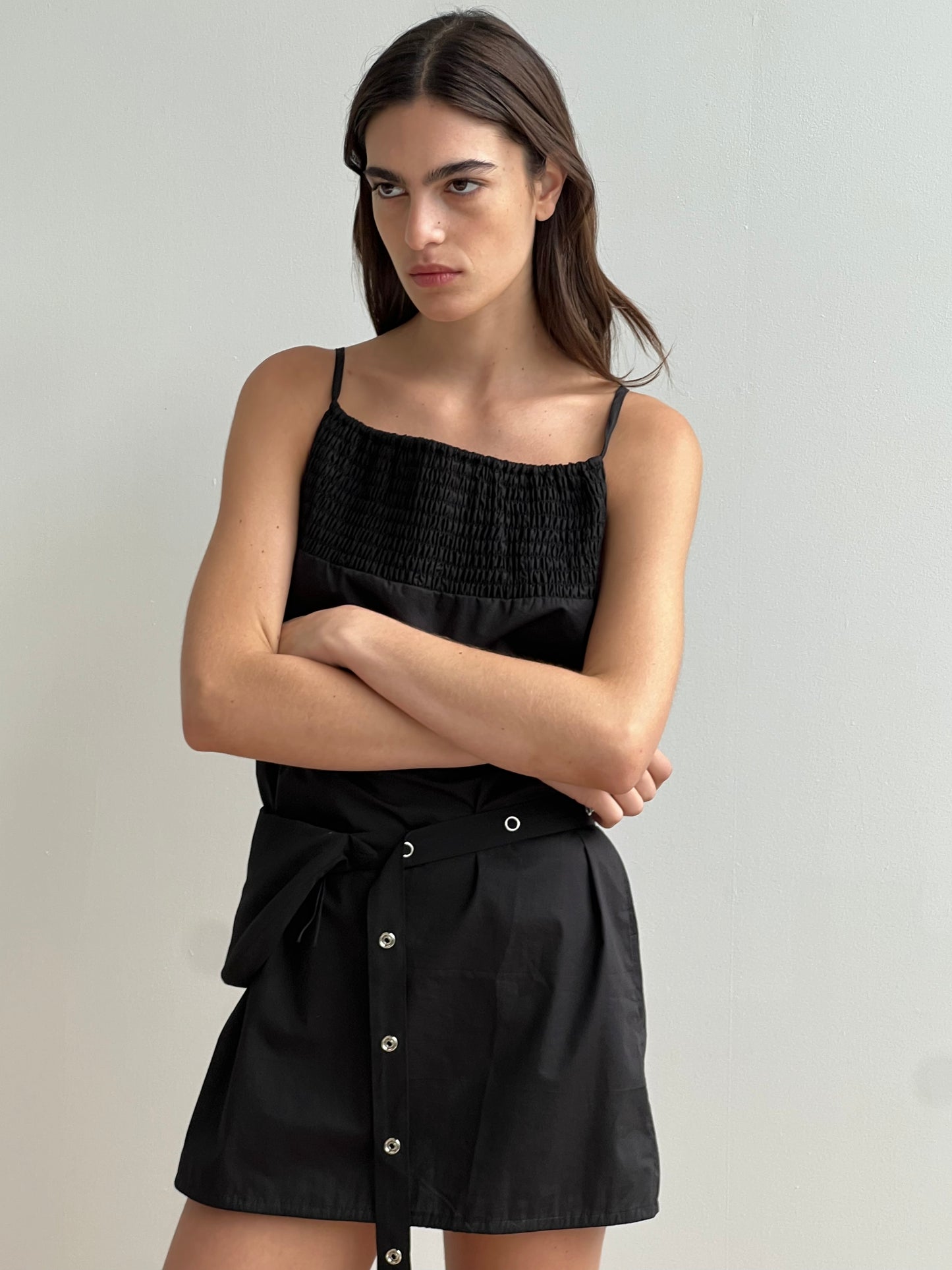 A woman with long brown hair stands against a plain light wall, wearing the Shirring Mini Dress - Black by Deiji Studios, featuring a smocked top and buttoned skirt. Her arms are crossed and she has a serious expression.