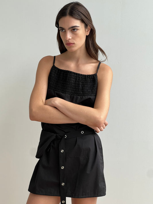 A woman with long brown hair stands against a plain light wall, wearing the Shirring Mini Dress - Black by Deiji Studios, featuring a smocked top and buttoned skirt. Her arms are crossed and she has a serious expression.