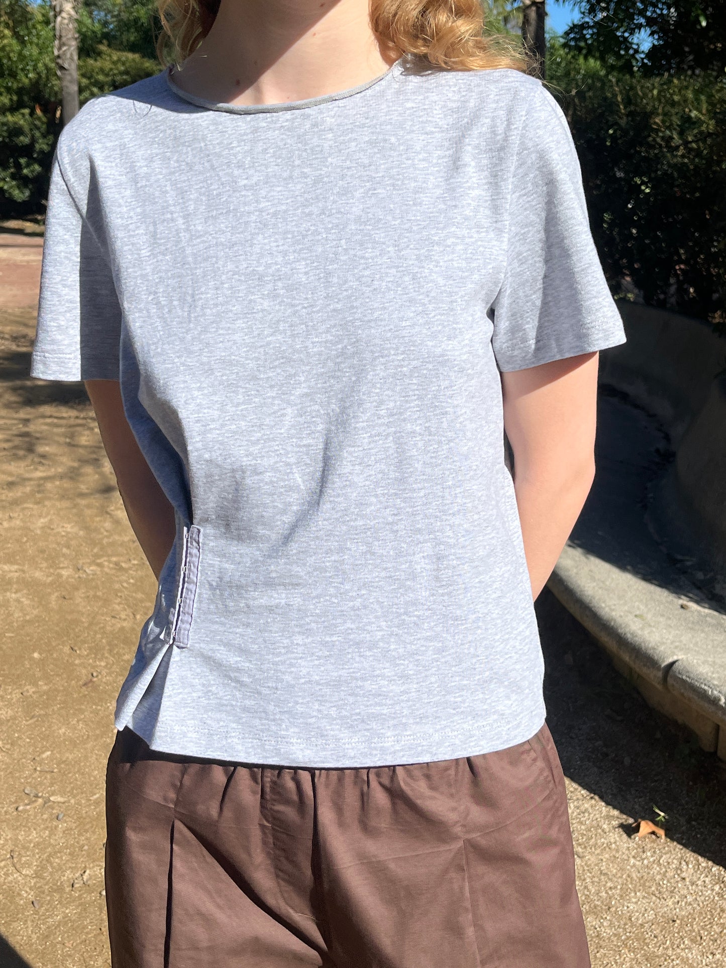 Outdoors, a person wears the Pinch Tee - Grey by Deiji Studios and loose brown pants as sunlight creates shadows; trees and a curved concrete bench are in the background.