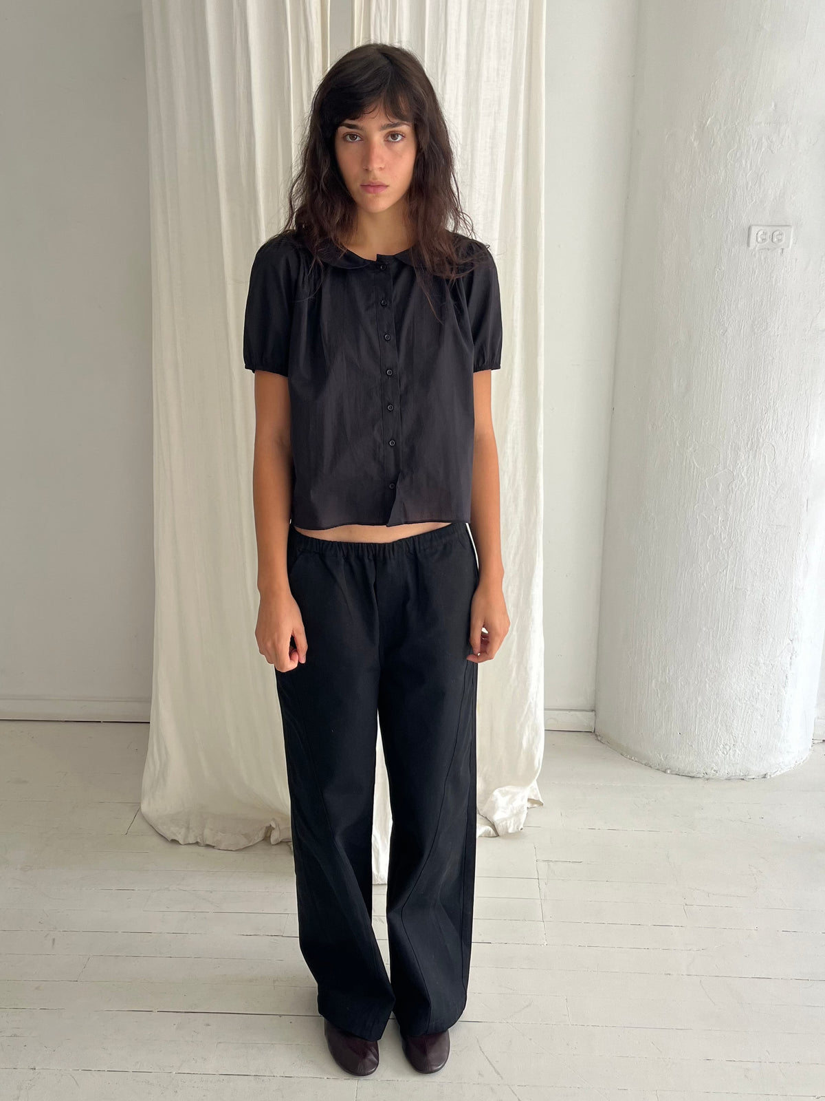 A woman with dark wavy hair stands against a white curtain, wearing the Curve Pant - Black by Deiji Studios, a softly structured pant with a relaxed fit and subtle curved leg, styled with the Petal Top in Black.