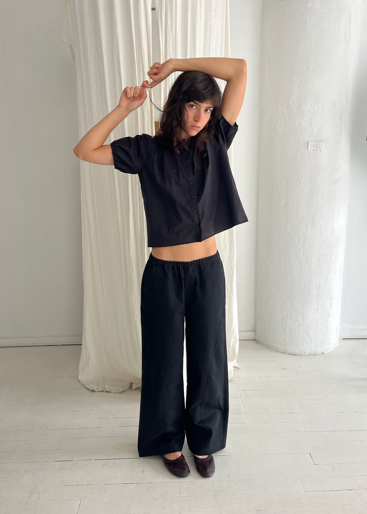 A woman stands against a white curtain with arms raised, wearing the Curve Pant - Black by Deiji Studios, a softly structured pant with a relaxed fit and subtle curved leg, styled with the Petal Top in Black.