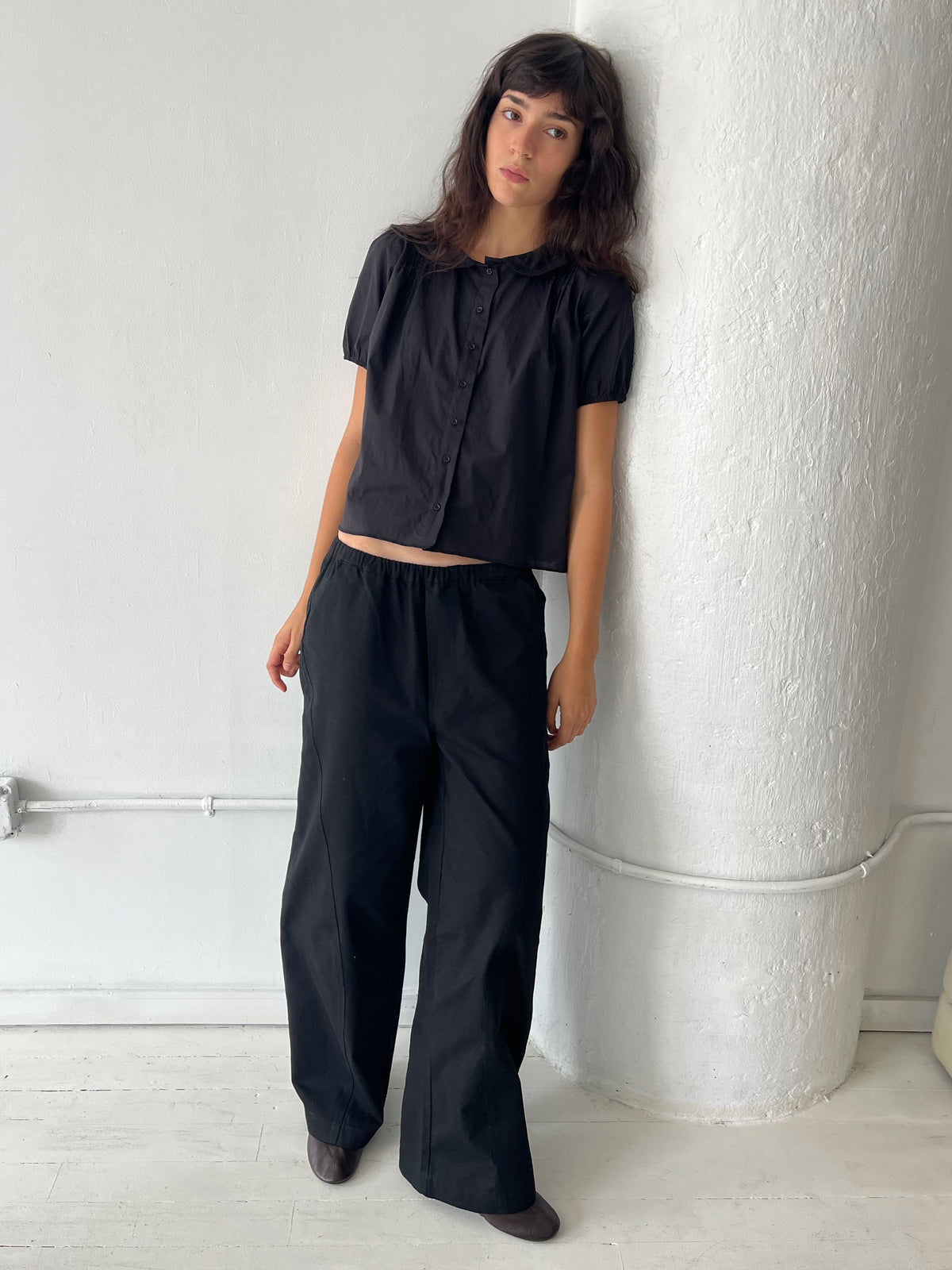 A woman leans against a white column, wearing the Curve Pant - Black by Deiji Studios, a softly structured pant with a relaxed fit and subtle curved leg, styled with the Petal Top in Black.