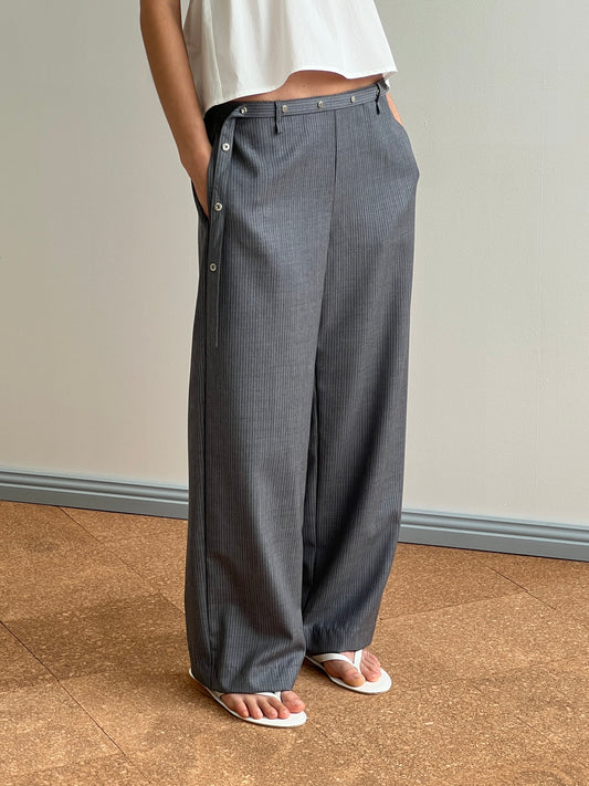 A person wearing the Wool Belt Pant - Grey Blue Stripe with side button details, a white cropped top, and white flip-flops stands indoors on a brown floor against a light wall.