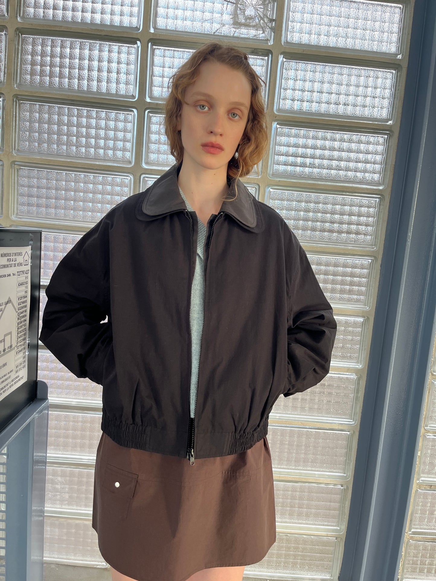 A person with short, wavy hair stands indoors by glass brick windows, wearing the Collared Zip Jacket - Black Contrast over a light shirt and a brown skirt with a front pocket, hands relaxed in the jacket’s pockets.