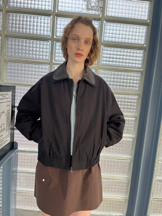A person with short, wavy hair stands indoors by glass block windows, wearing the Collared Zip Jacket - Black Contrast over a light top and brown skirt, looking directly at the camera with a neutral expression.